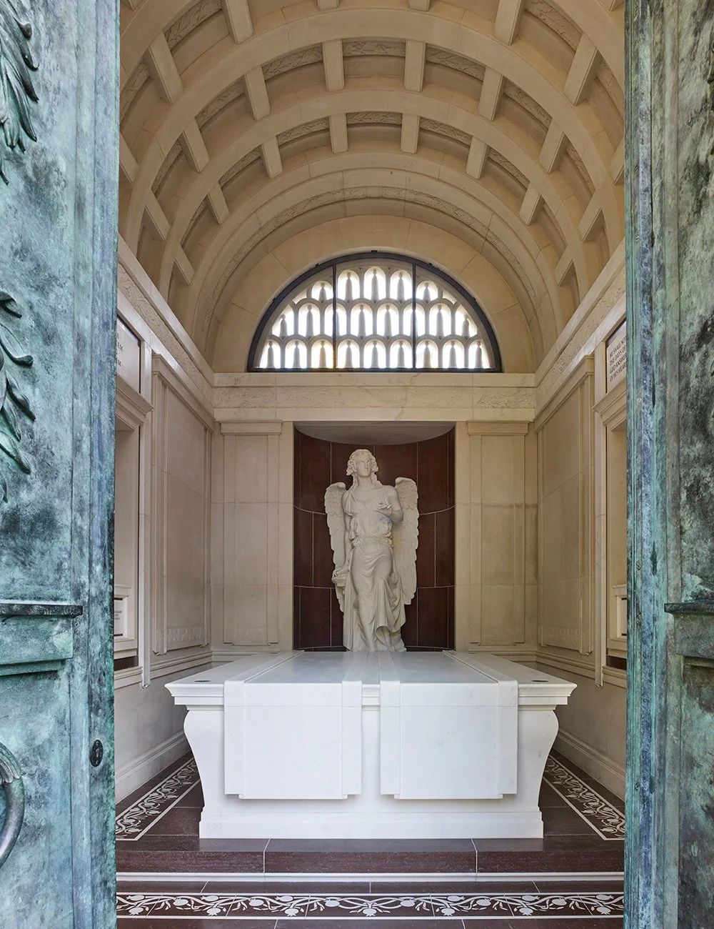 Interior of a classical mausoleum featuring a marble angel sculpture beneath a coffered stone vault and traditional funerary detailing.