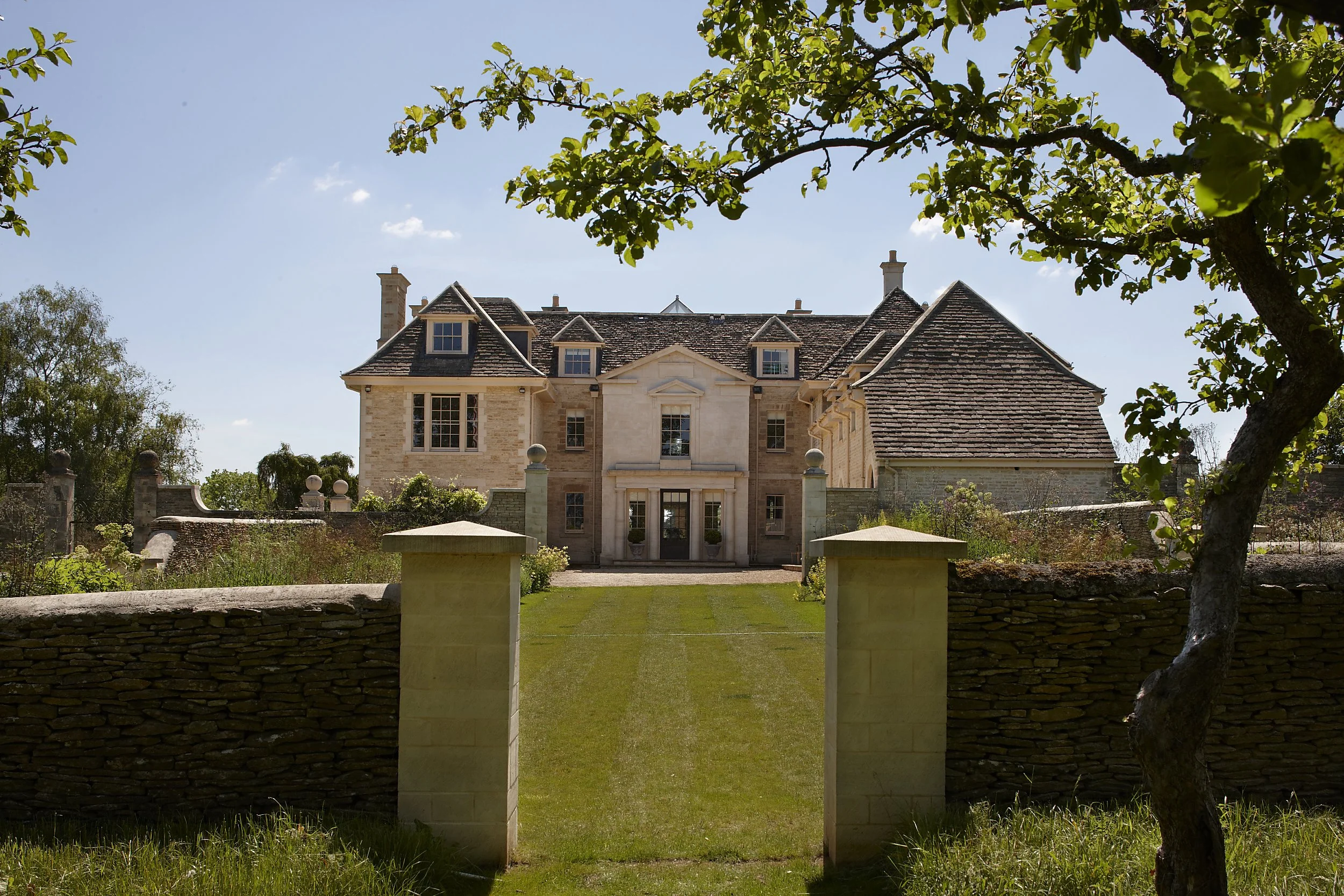 Tree-framed view of a traditional Cotswolds country house with formal lawn approach and classical limestone architecture by Craig Hamilton Architects
