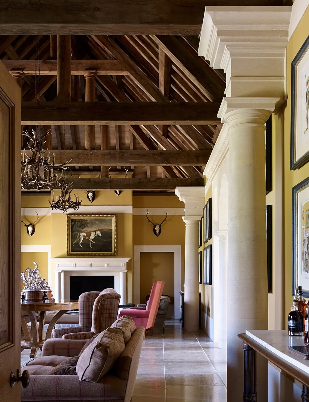 View through the great hall of the Shooting Lodge in Gloucestershire featuring exposed timber roof trusses, classical columns and a formal fireplace.