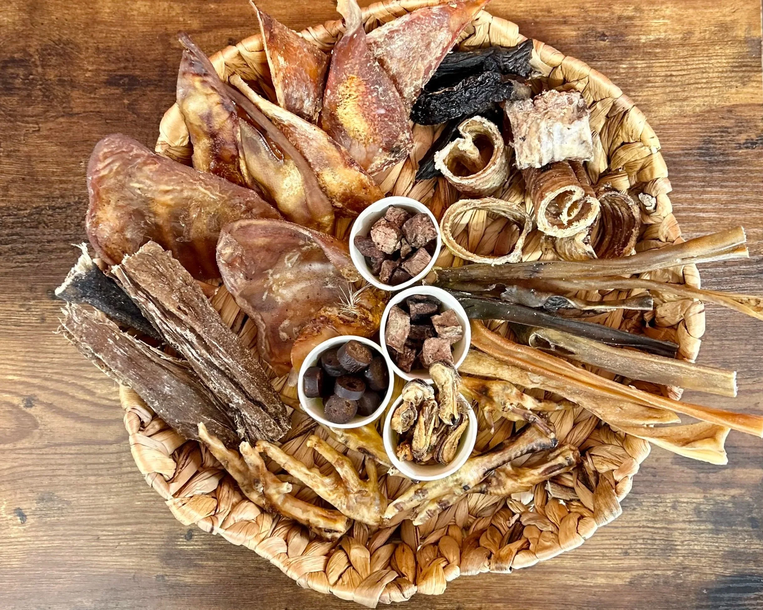 A round woven basket filled with various natural dog treats, and seafood, placed on a wooden surface.