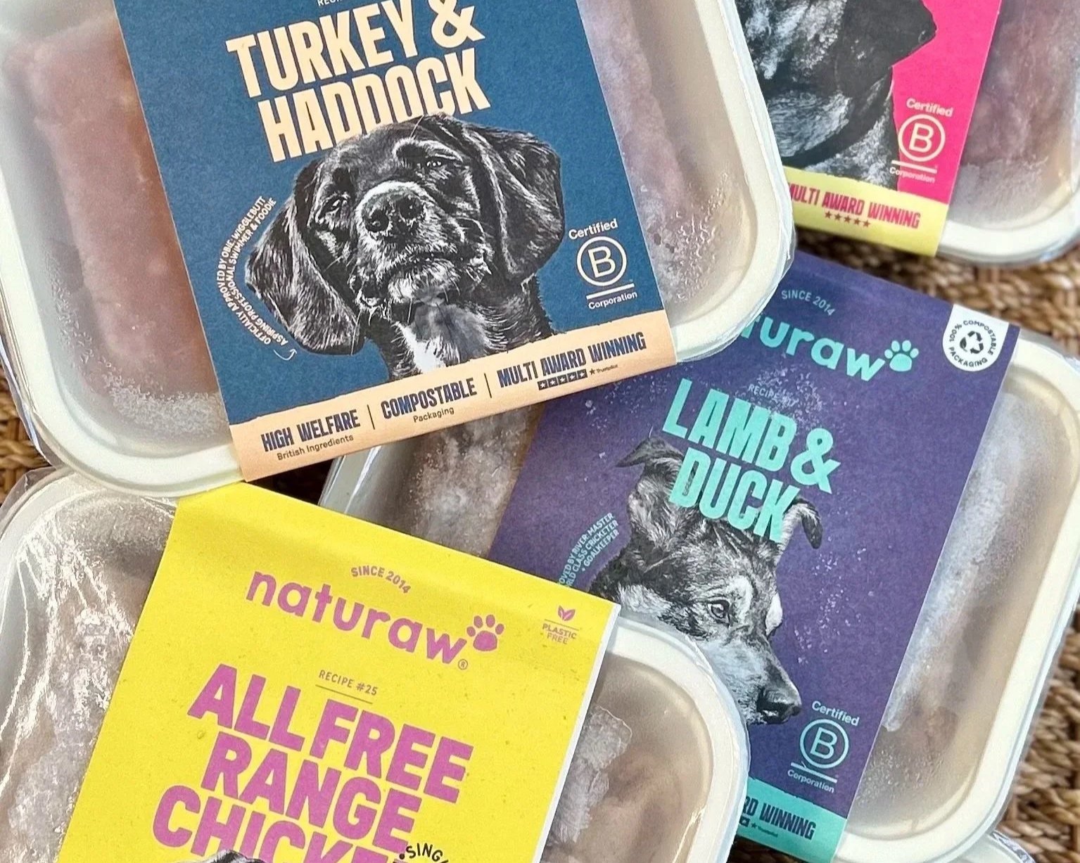 Several packages of raw pet food with colorful labels featuring illustrations of dogs. The labels display product names like 'Turkey & Haddock', 'Lamb & Duck', and 'All Free Ranged Chicken', with various certifications and product highlights.