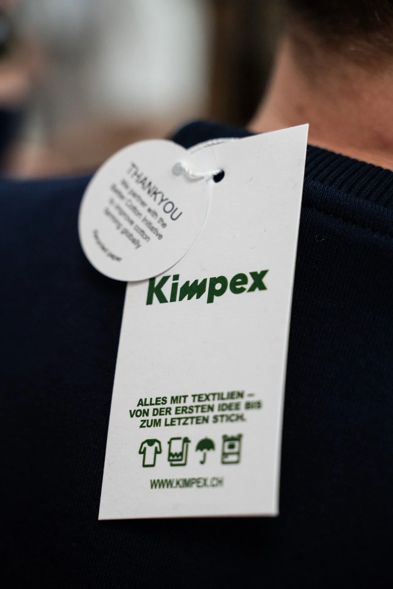 Kleidungsetikett mit dem Logo 'Kimpex' und Text auf Deutsch, das Textilien und Pflegehinweise beschreibt.