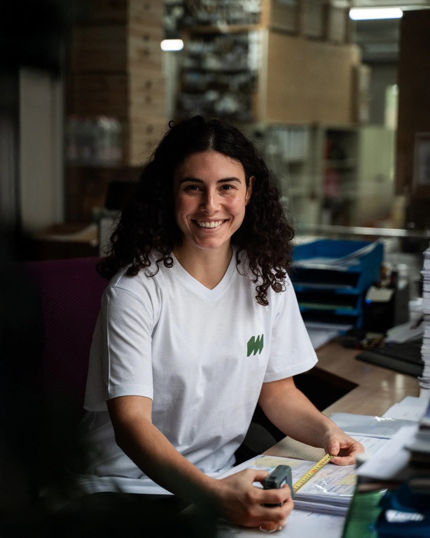 Eine junge Frau sitzt an einem Schreibtisch, lächelt und hält ein Maßband in der Hand, umgeben von Akten und Papieren, im Hintergrund eine Holzwand und Regale.