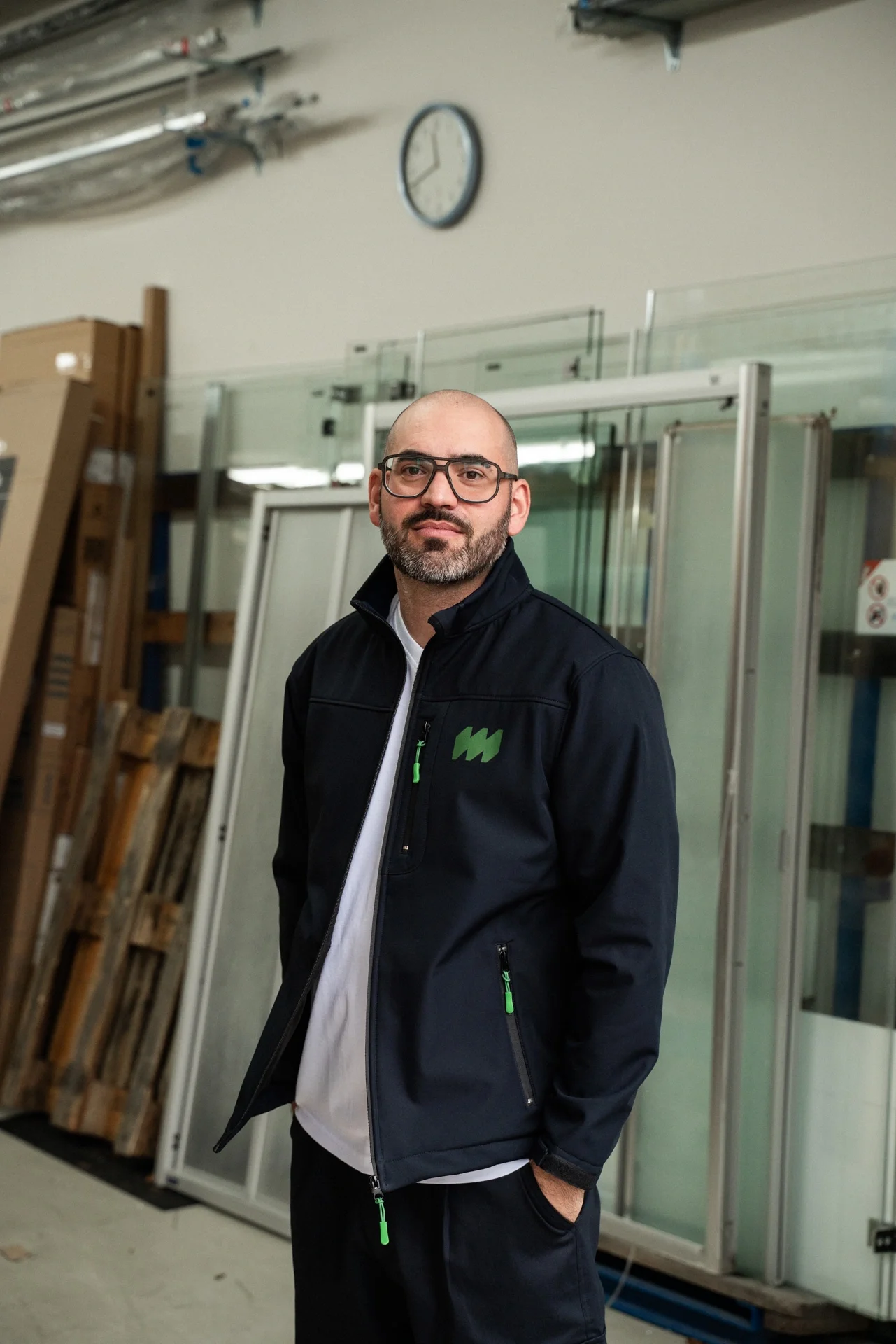 Ein Mann mit Brille und Bart trägt eine schwarze Jacke mit grünen Details. Er steht in einem Raum mit Industrieausrüstung und Glasfenstern, im Hintergrund sind Bauelemente und eine Uhr an der Wand.