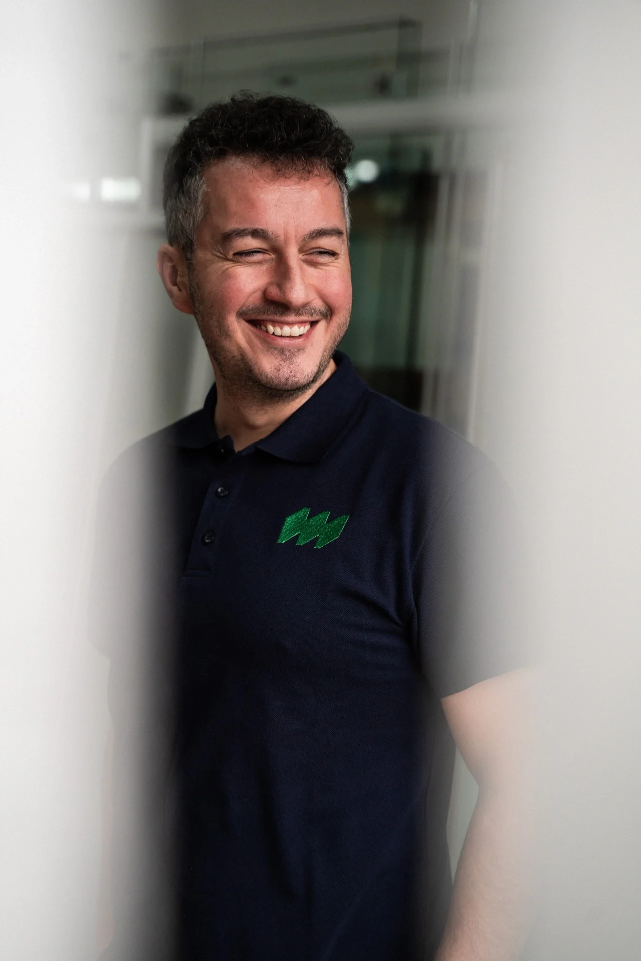 Lächelnder Mann trägt dunkelblaues Polo-Shirt mit grüner Logo auf der Brust.