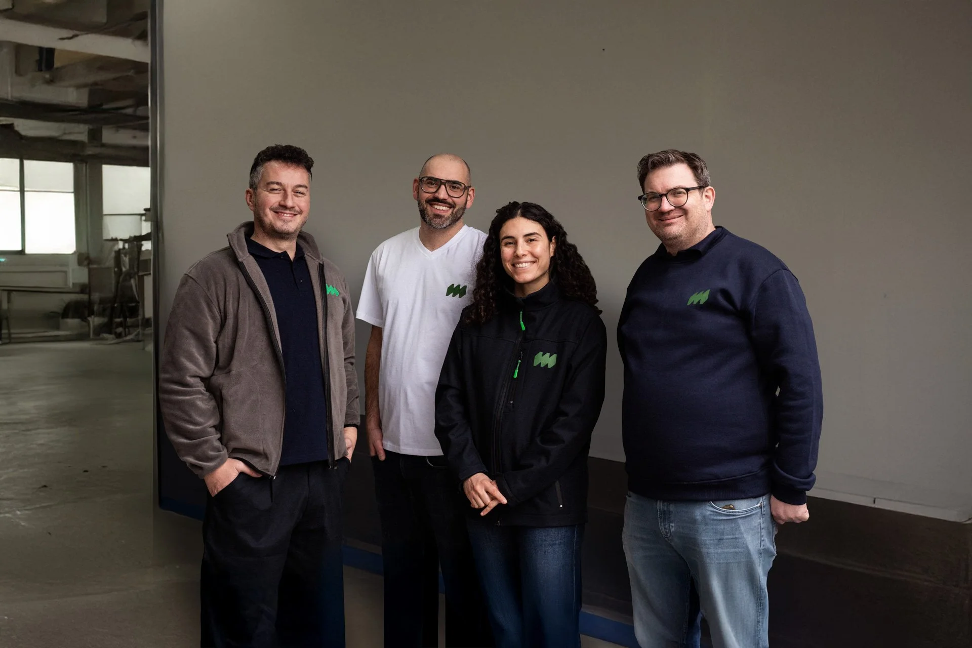 Vier Menschen stehen vor einer grauen Wand und lachen in die Kamera. Zwei Männer und eine Frau tragen dunkle Kleidung mit einem grünen Logo, eine weitere Person trägt ein weißes T-Shirt.