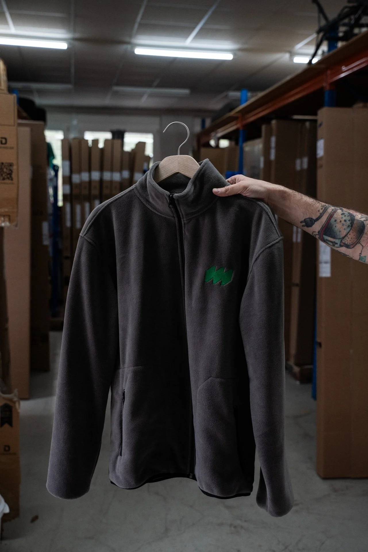 Eine schwarze Fleecejacke mit grünem Logo, die an einer Kleiderstange in einem Lager hängt, von einer Person mit tätowiertem Arm gehalten.