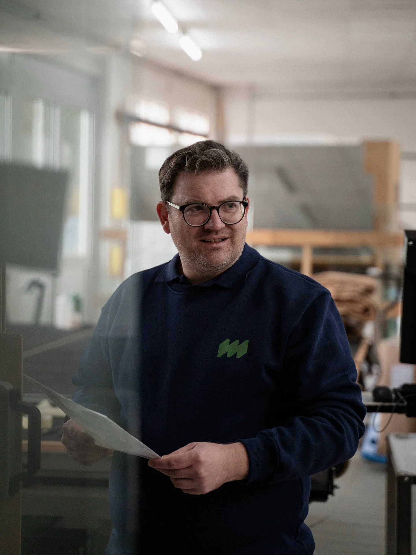 Ein Mann mit Brille sitzt in einem Büro und liest Dokumente, im Hintergrund sind Büroregale und offene Fenster sichtbar.