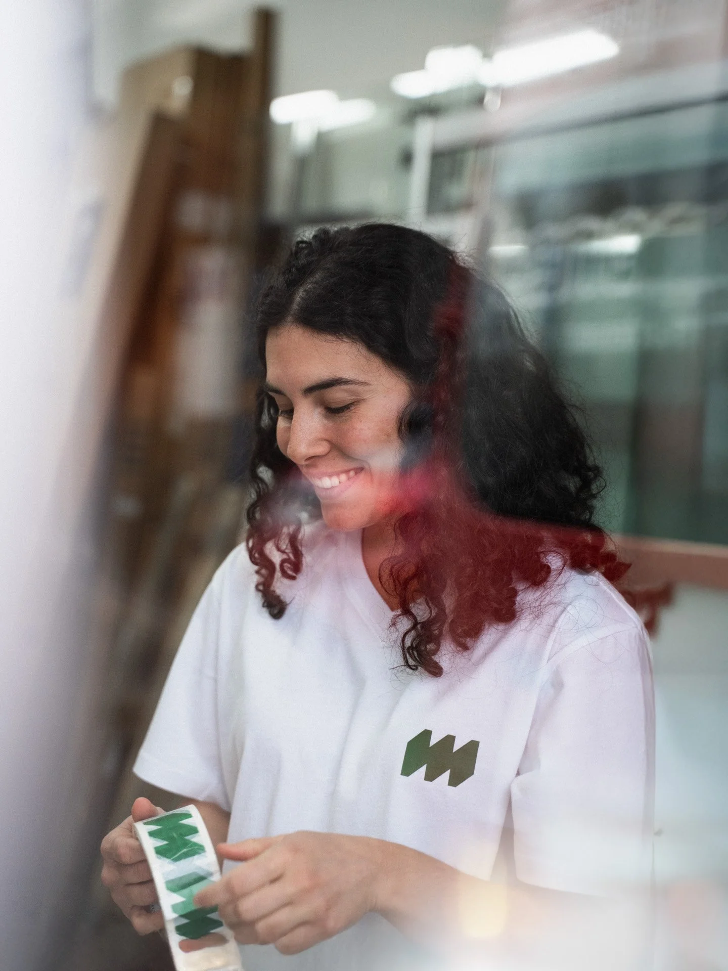 Junge Frau mit dunklen, lockigen Haaren trägt ein weisses T-Shirt mit einem grünen Logo, lächelt und hält eine Etikette in der Hand, durch ein Fenster fotografiert.