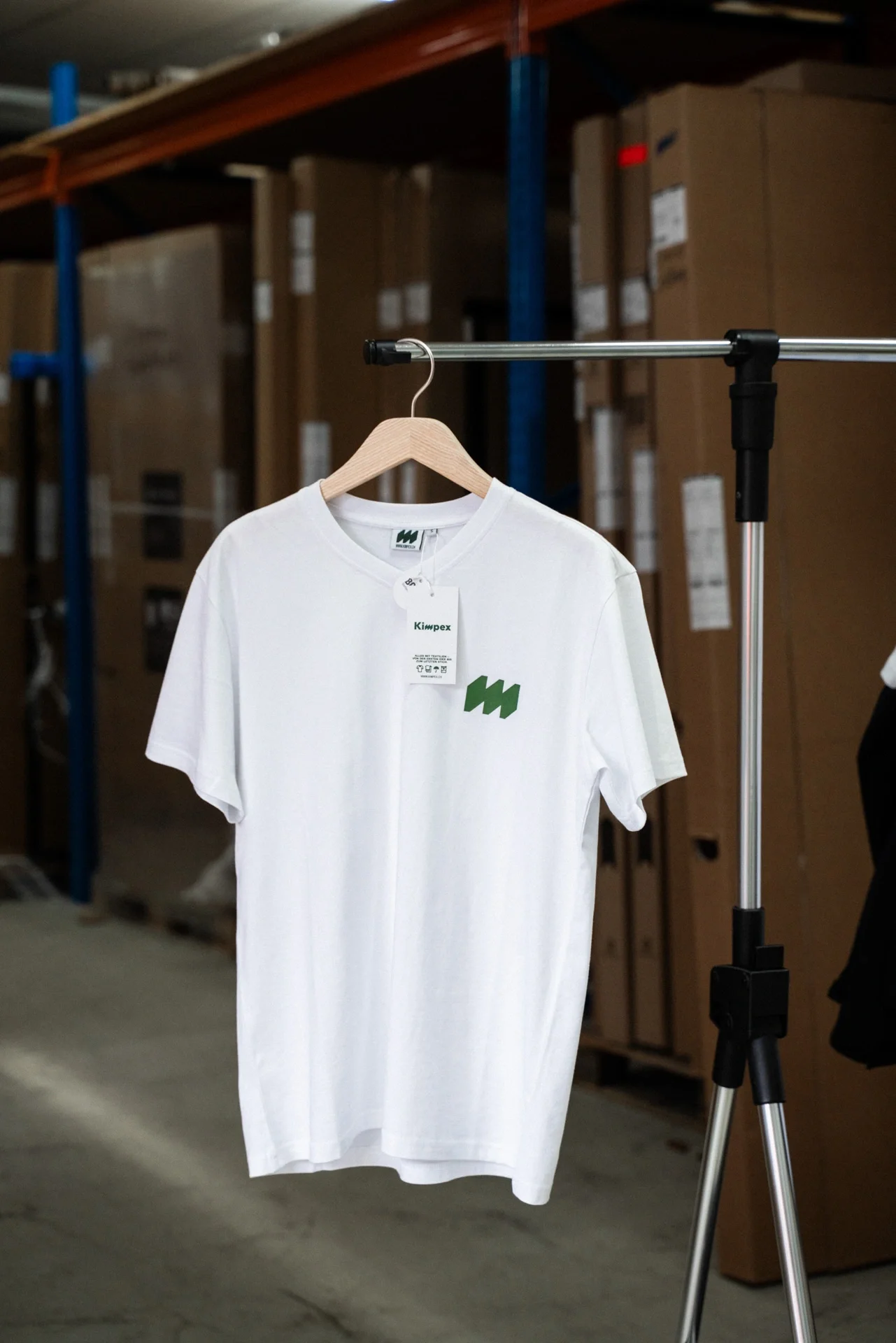 Ein weisses T-Shirt mit einem grünen Logo hängt an einem Holzkleiderbügel an einer Kleiderstange in einem Lagerraum mit Umzugskartons im Hintergrund.