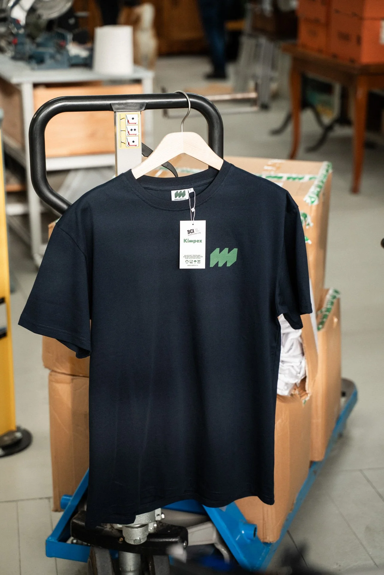 Ein schwarzes T-Shirt mit einem grünen Logo hängt an einem Kleiderbügel an einem Handwagen in einem Lager oder einer Werkstatt.