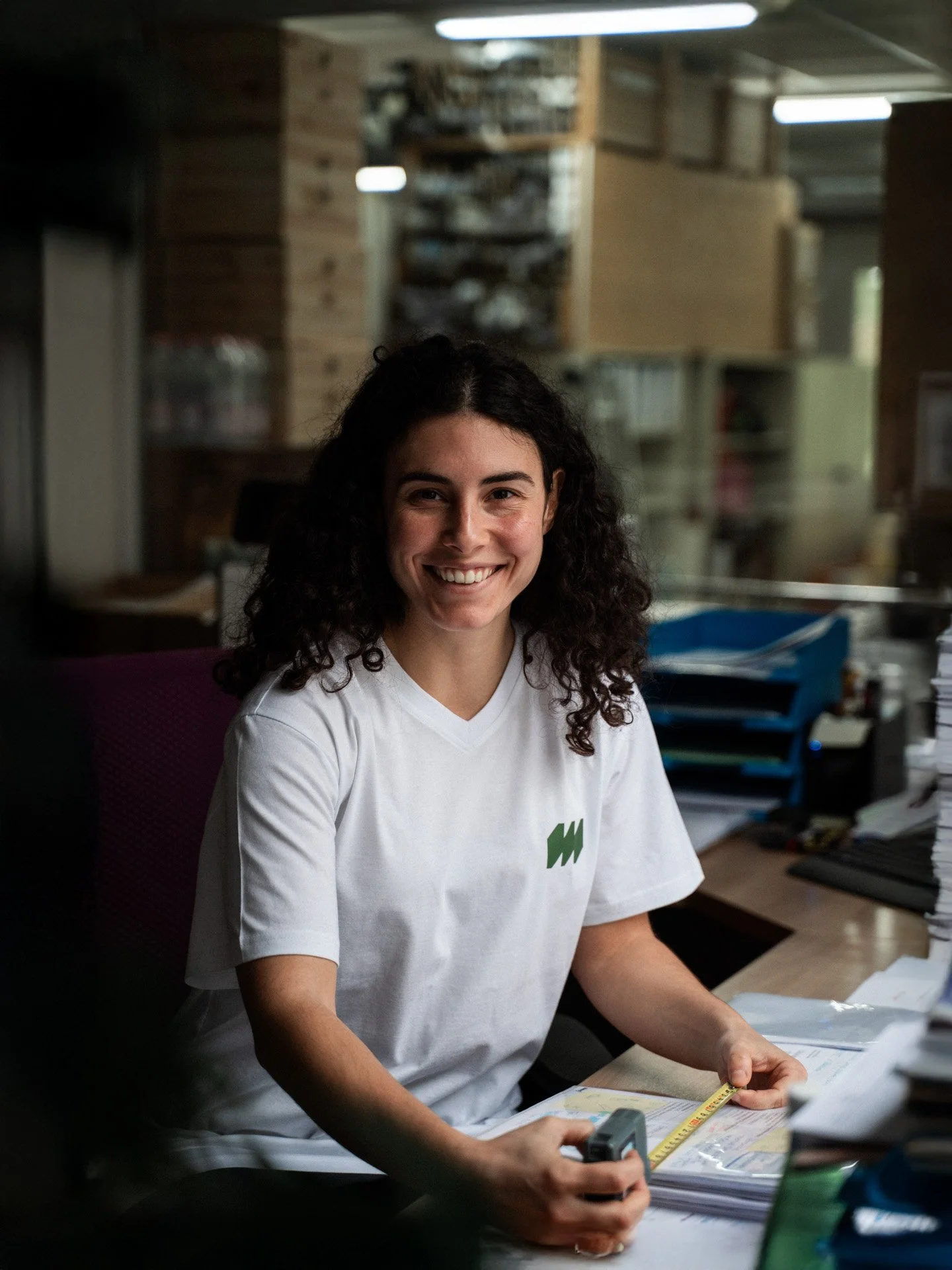 Lächelnde junge Frau mit lockigem schwarzen Haar arbeitet an einem Schreibtisch mit Akten und einem Massband in einem Büro mit Holzregalen im Hintergrund.