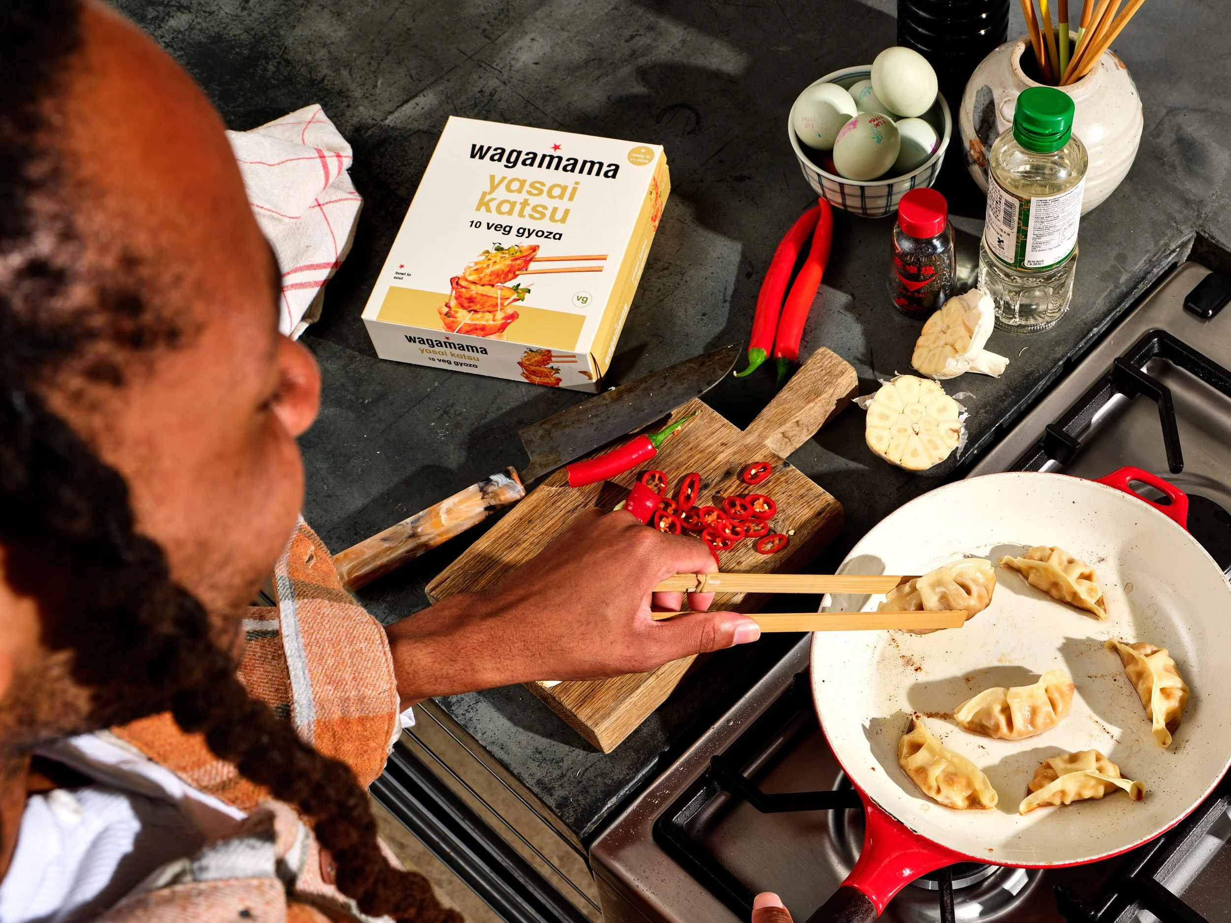 Person preparing gyoza dumplings in a frying pan on a stove, with chili peppers and cooking ingredients on the countertop, and a box of plant-based veggie gyoza nearby.