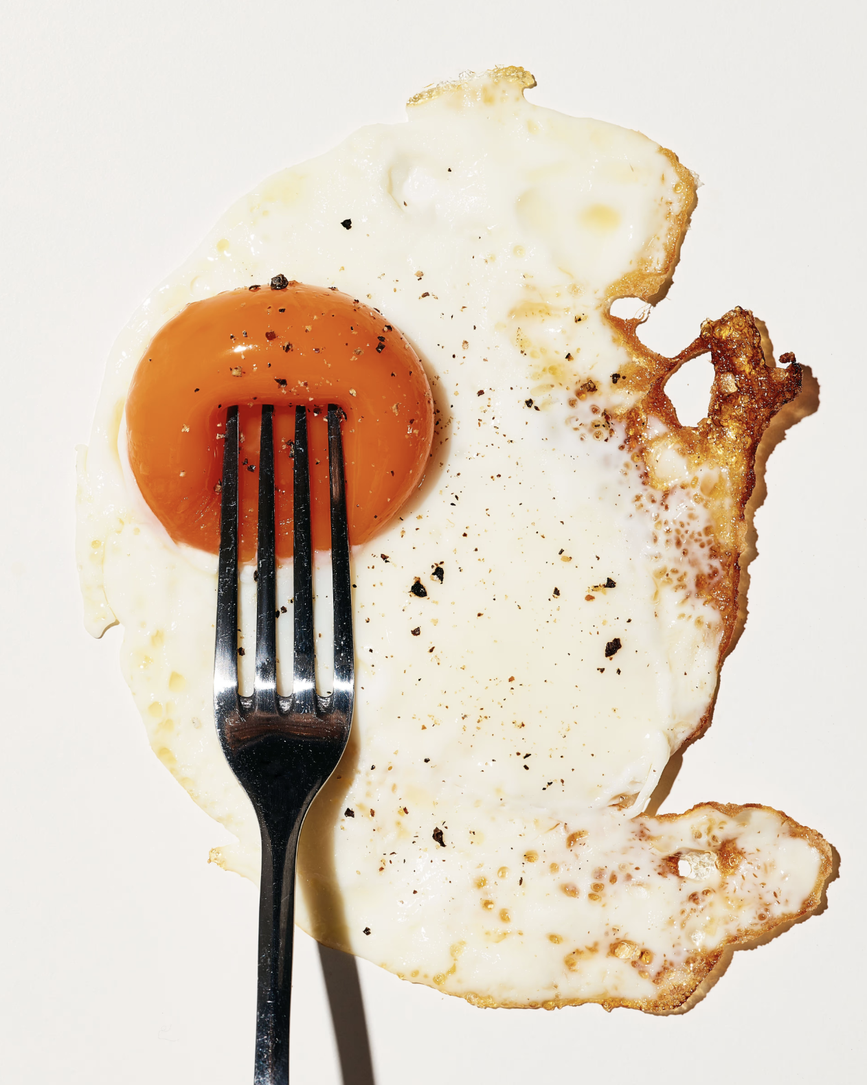 Fried egg with a runny yolk on a white background, topped with black pepper and salt, with a fork piercing the yolk.