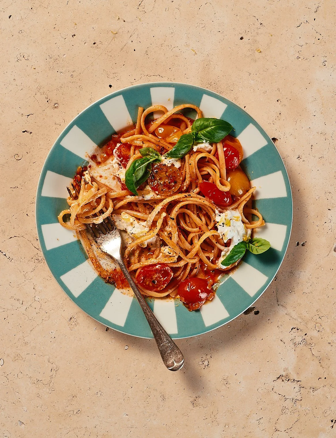 A plate of spaghetti pasta with cherry tomatoes, fresh basil leaves, and a sauce, on a textured beige surface.