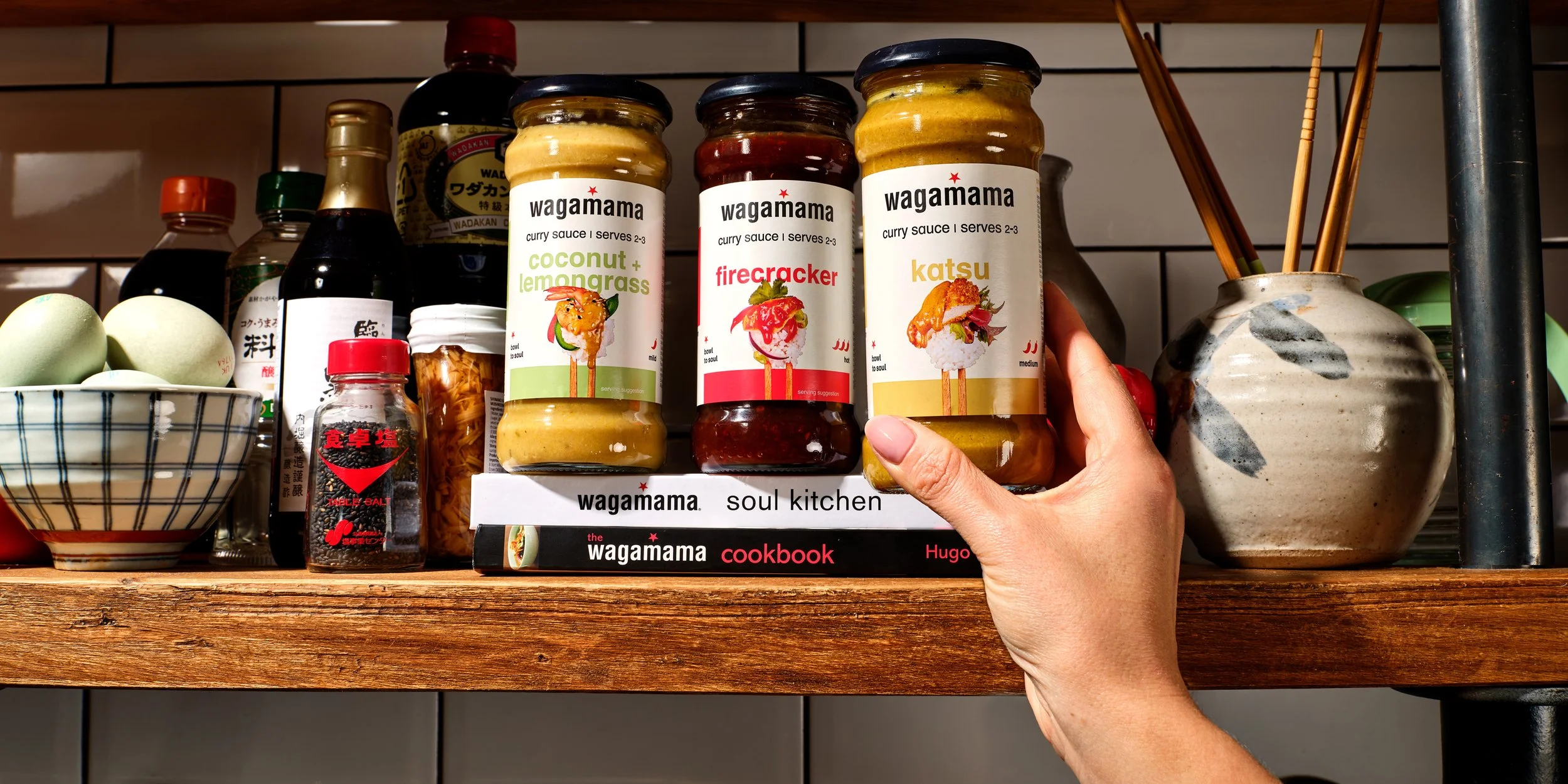 Hand holding a jar of Wagamama curry sauce in front of other curry sauces on a store shelf, with a bowl of eggs and various bottles in the background.
