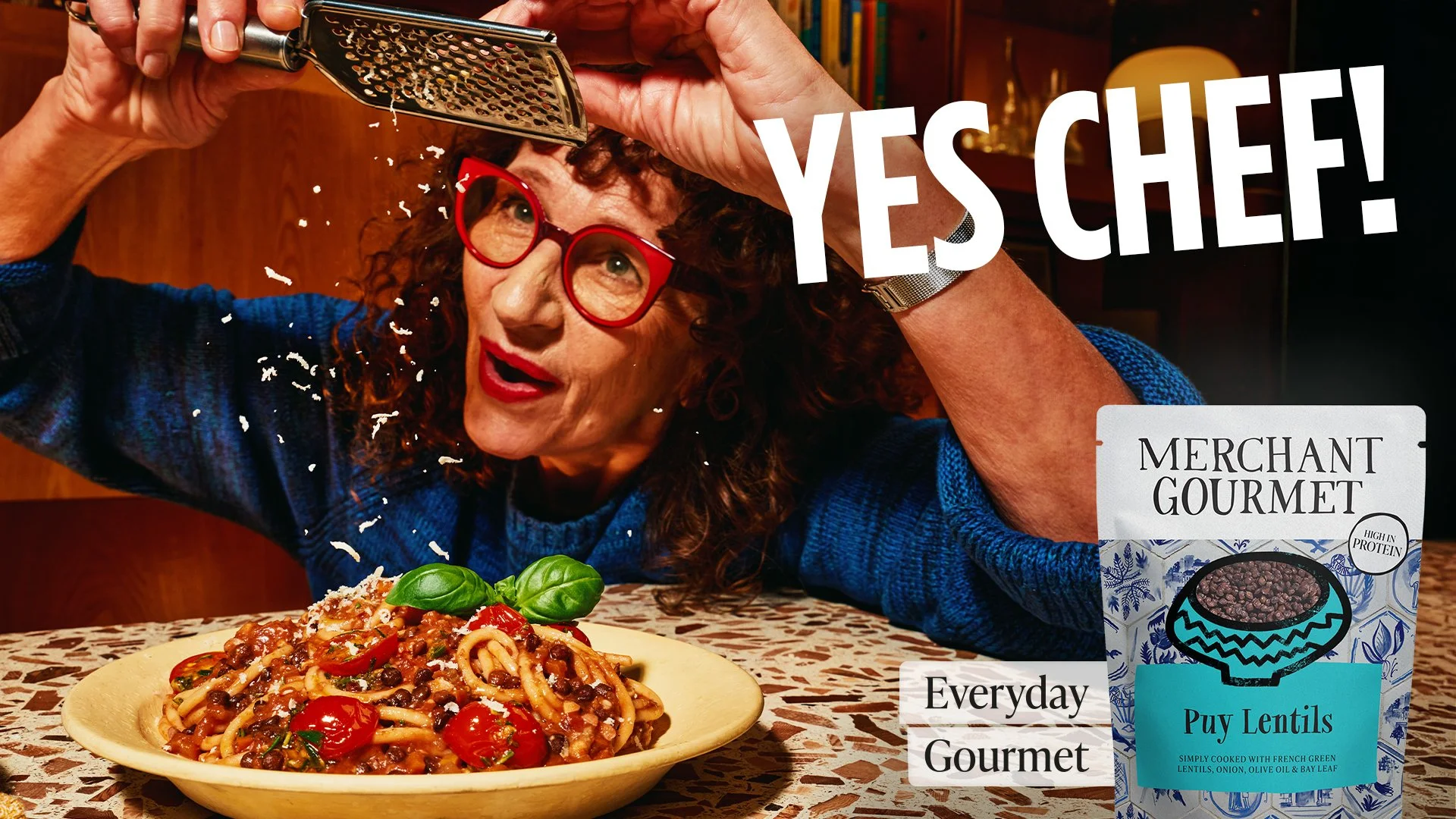 A woman with curly hair and red glasses is grating cheese over a plate of pasta with cherry tomatoes and basil, on a table with a patterned tablecloth. There is a package of lentils on the right side of the image.