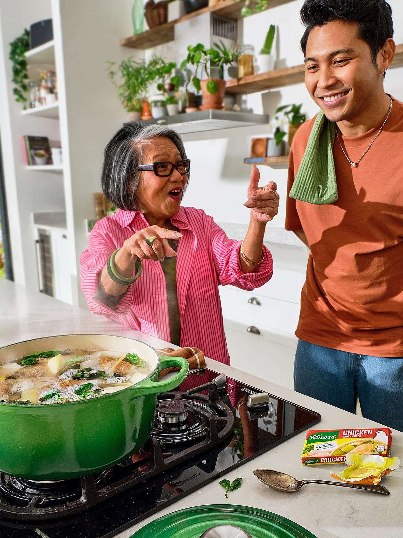 A woman and a man in a kitchen smiling and talking to each other while cooking. On the counter, there is a green pot with soup, and a spoon. Grocery items include a box of chicken broth cubes. The background shows shelves with plants and kitchen uten