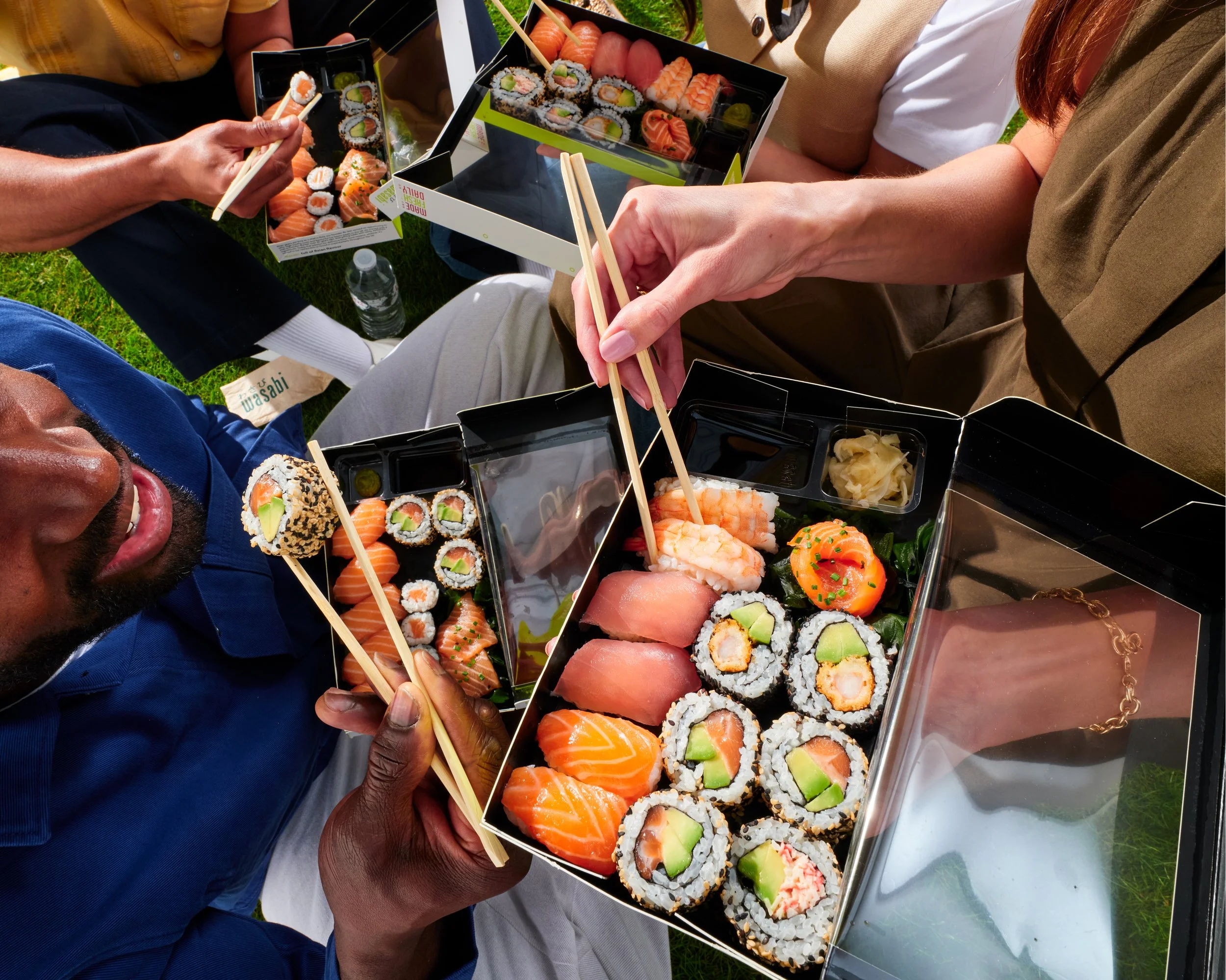 People enjoying a variety of Wasabi sushi rolls outdoors on a picnic table. The sushi includes options with salmon, shrimp, avocado, and cucumber, with some rolls topped with fish slices or garnishes, and a small bottle of water visible on the table.
