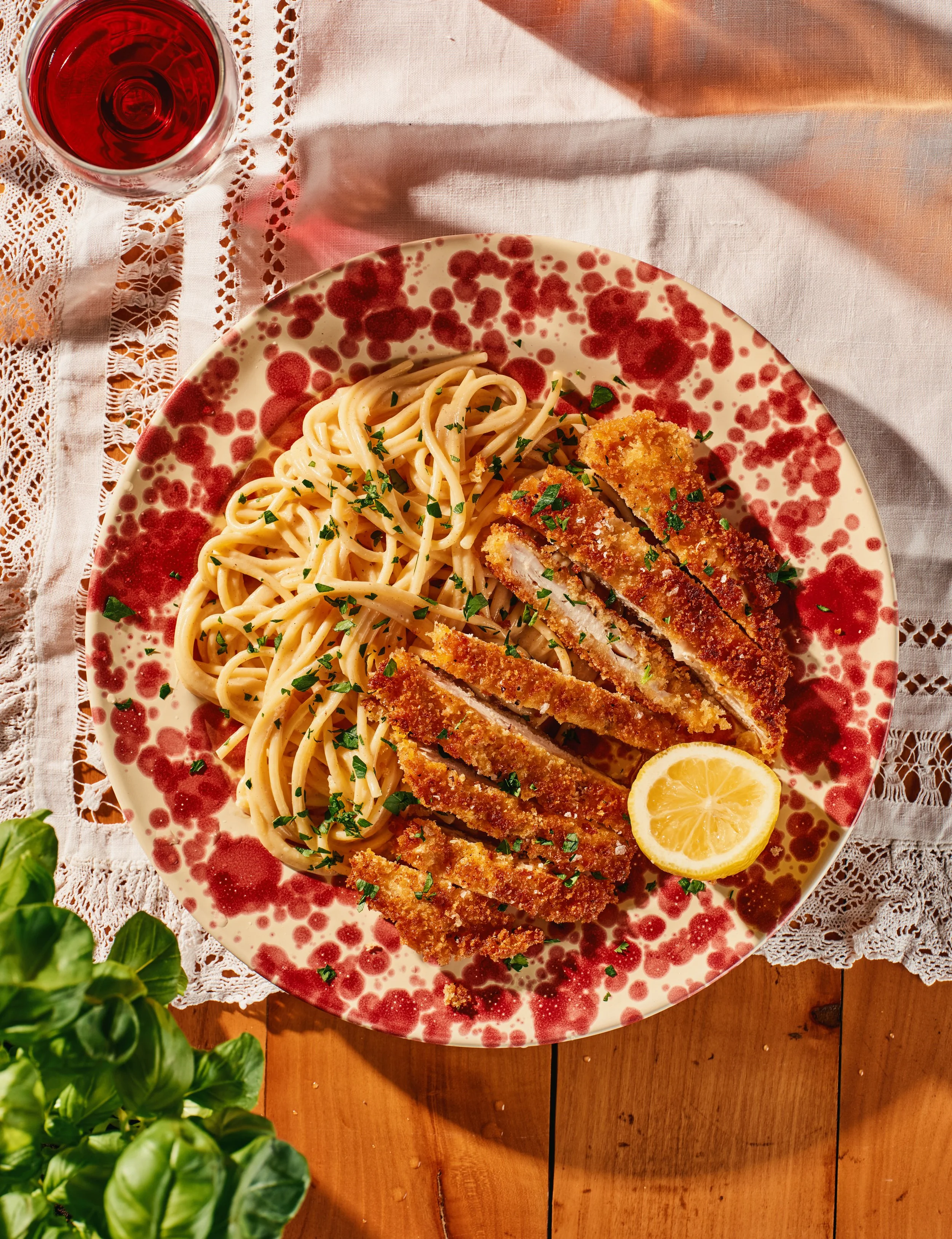 A plate with spaghetti and breaded chicken cutlets garnished with chopped herbs, with a lemon wedge on the side, on a rustic wooden table. A glass of red wine is in the top left corner, and the plate is on a white lace tablecloth with pink accents.
