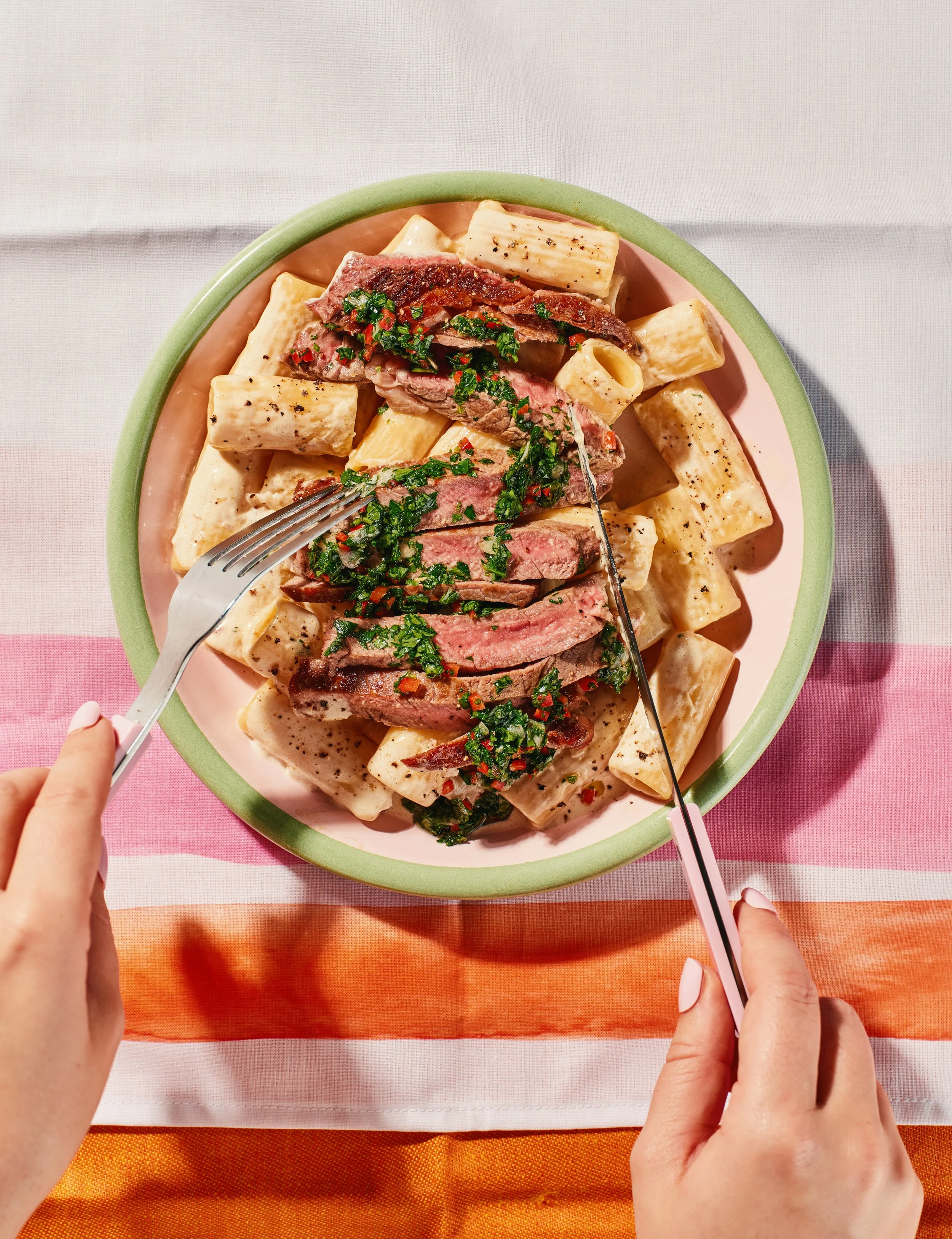 Overhead view of a pink plate filled with rigatoni pasta and sliced steak, topped with chopped parsley and red pepper flakes, on a multicolored tablecloth.