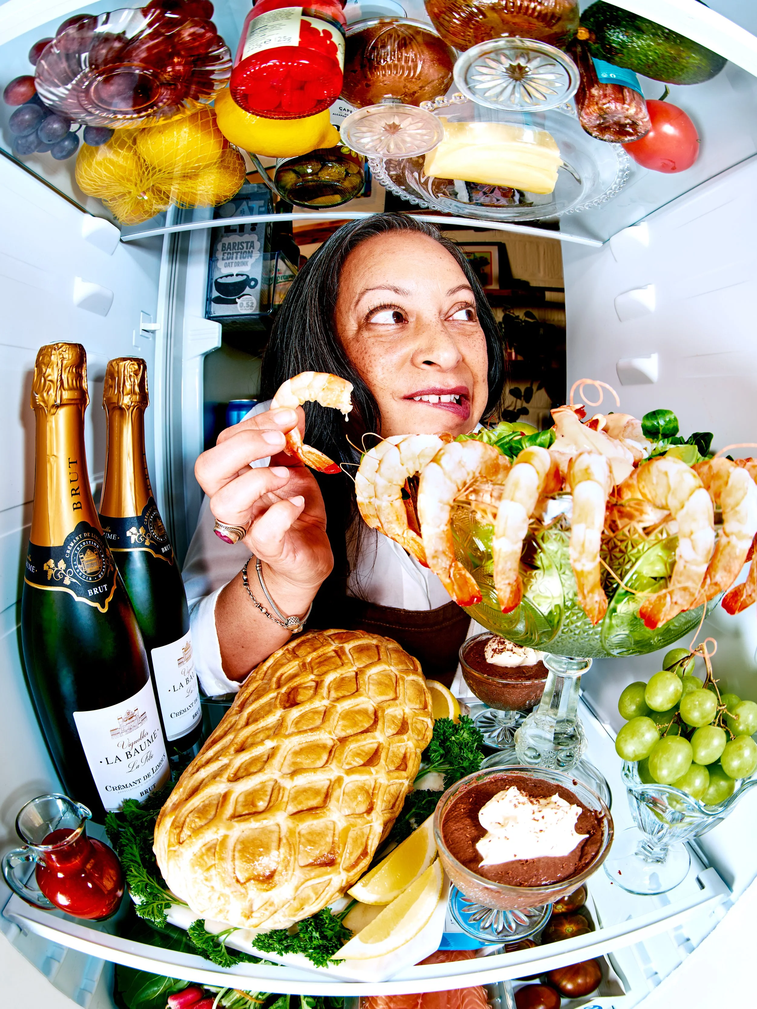 Inside a refrigerator with various food items including a woman looking inside, bottles of champagne, a loaf of bread, grapes, lemon slices, shrimp, bowls of dessert, and other fresh produce.
