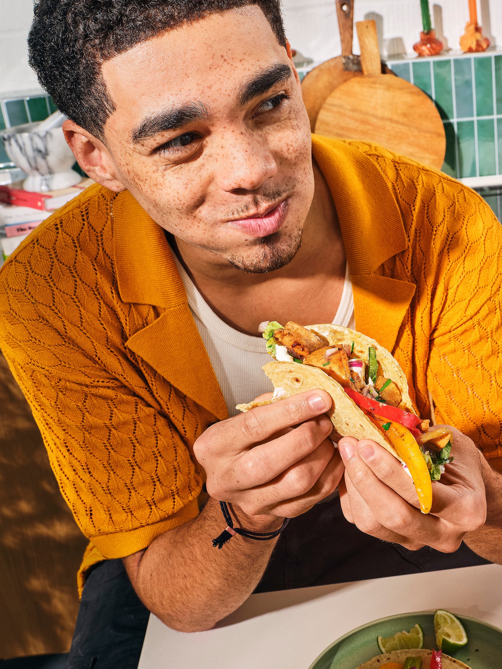 A man with short curly dark hair, brown skin, and freckles, wearing a yellow textured jacket, is holding and about to eat a taco with various fillings, including chopped vegetables and possibly meat inside a soft tortilla. In the background, there ar