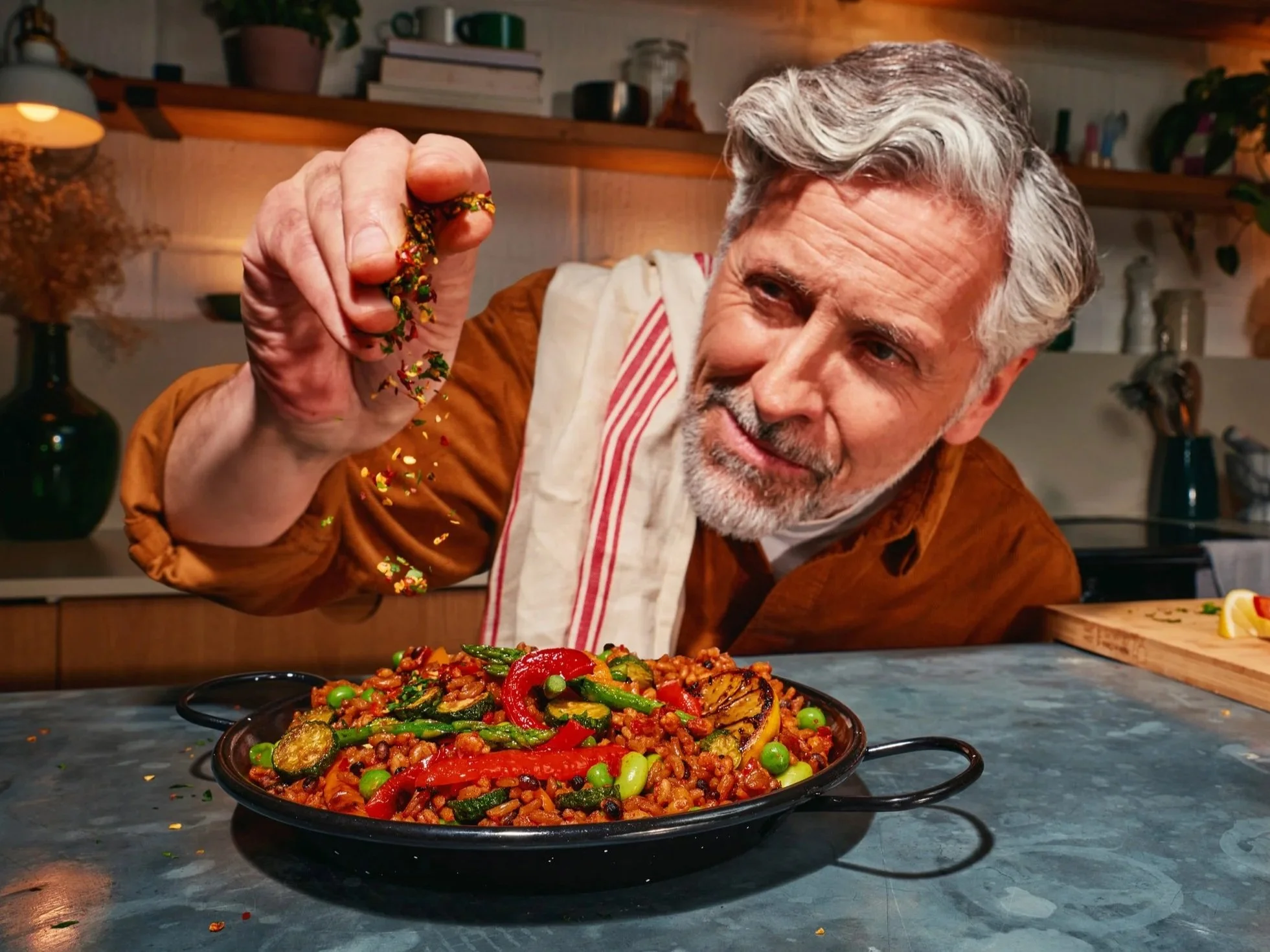 An elderly man with gray hair and a beard, wearing a brown shirt and a striped apron, is standing in a kitchen, holding colorful spices or herbs over a large skillet filled with a vegetable and grain dish that includes red peppers, green beans, and z
