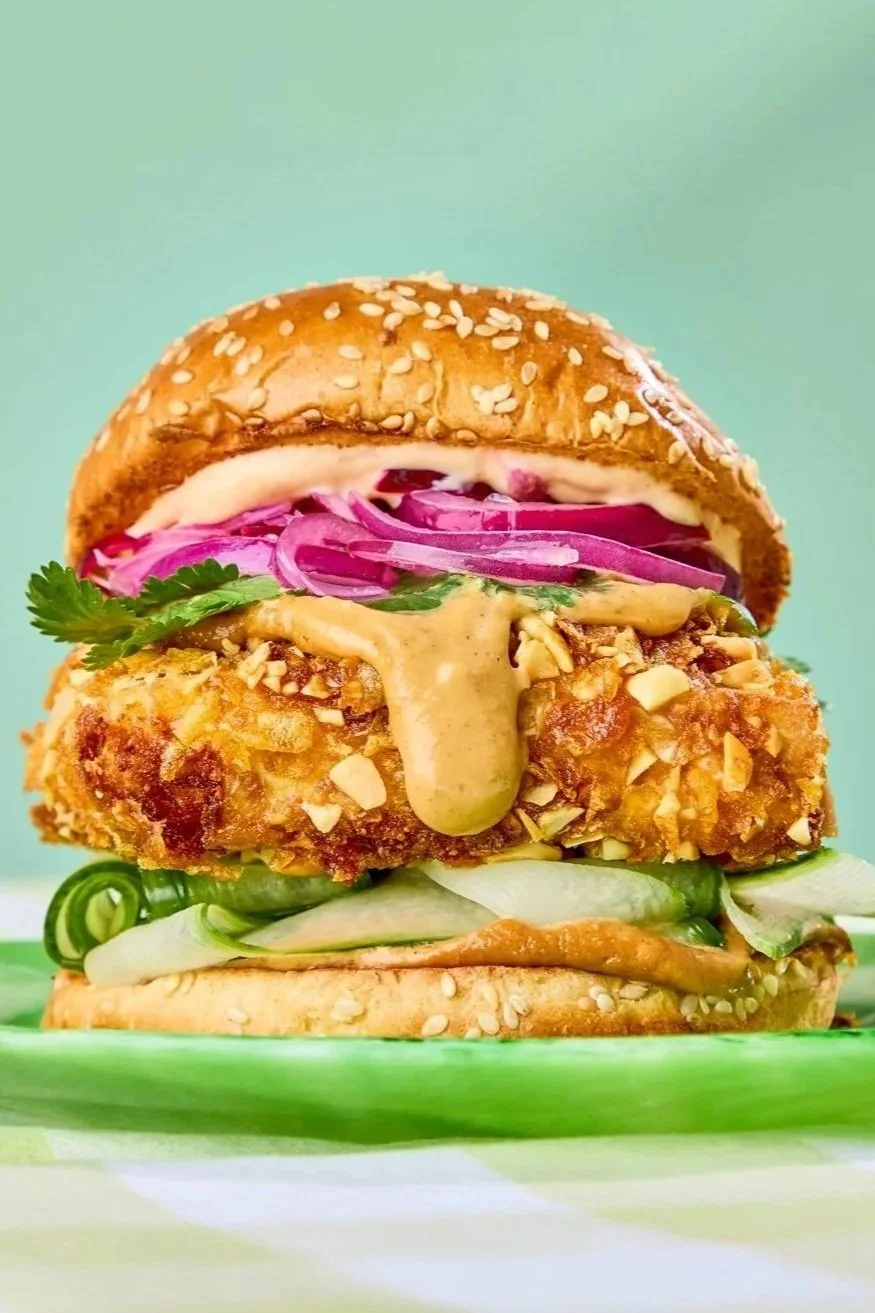Close-up of a fried fish sandwich with pickles, diced onions, cilantro, purple cabbage slaw, and sauce on a sesame seed bun, on a green plate with a green background.