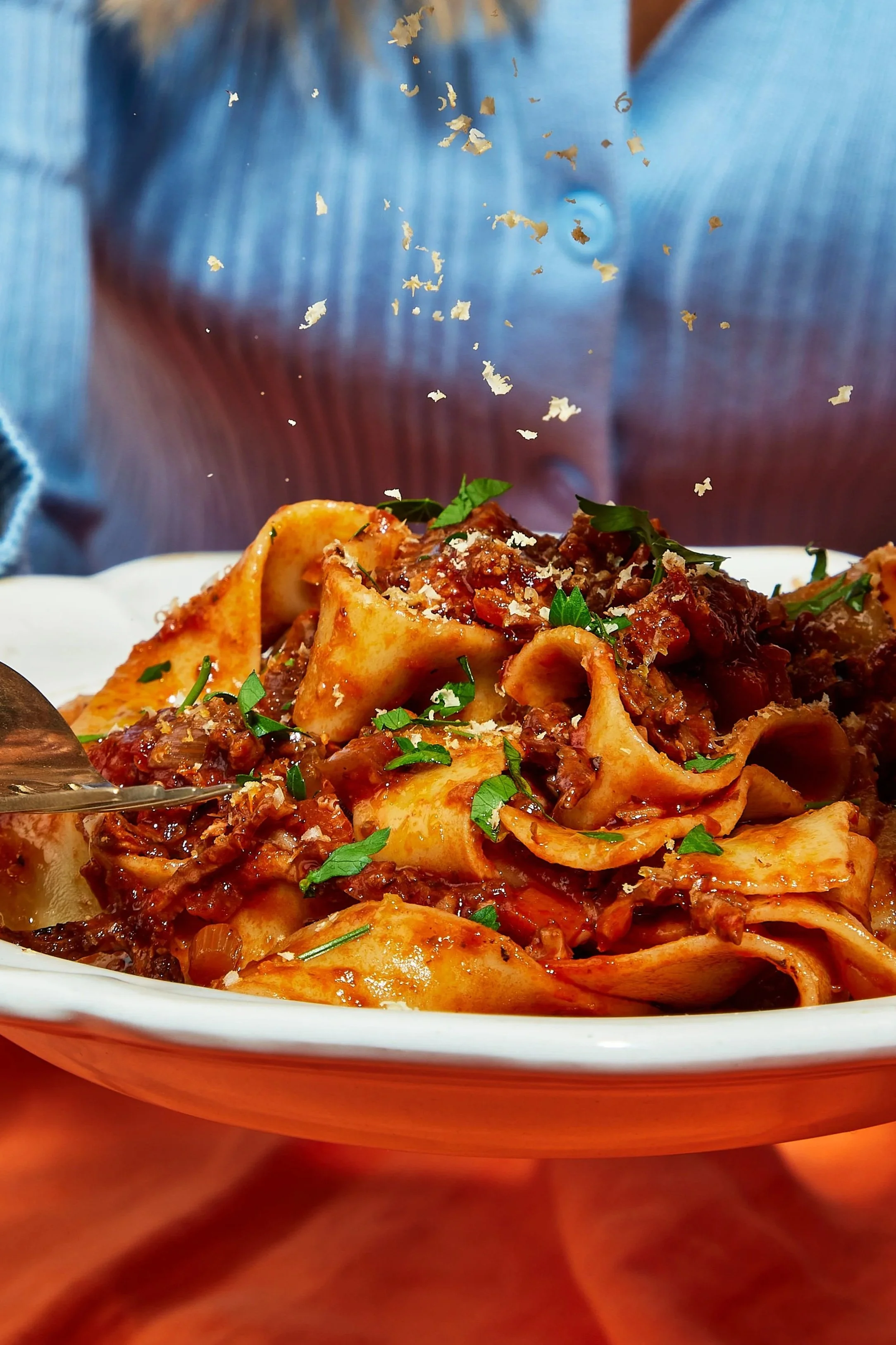 Close-up of a plate of cooked pasta with a tomato-based meat sauce, sprinkled with chopped herbs and grated cheese, with a person's face partially visible in the background.