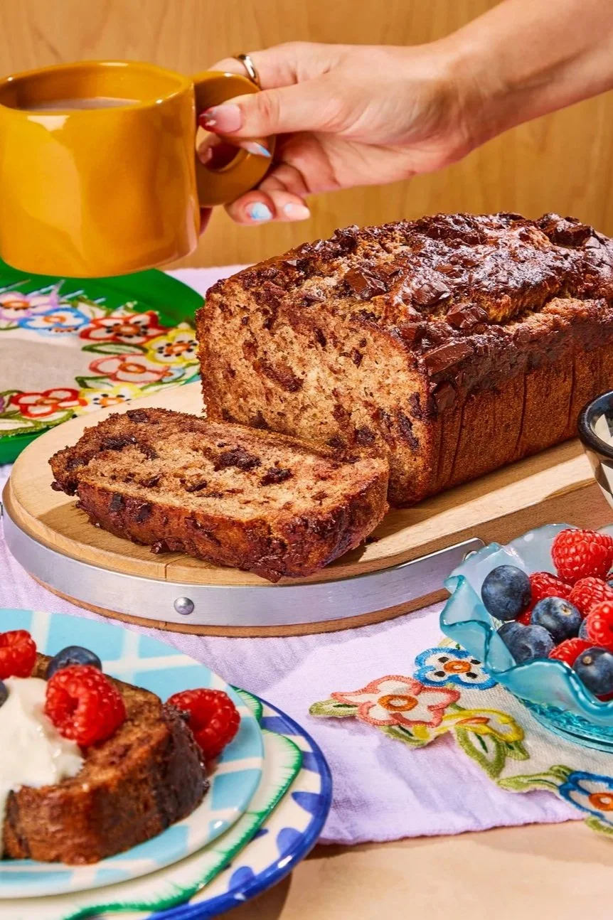 A loaf of banana bread on a wooden cutting board with one slice cut and placed on a colorful plate with berries, a person holding a mustard yellow mug over the bread, a colorful embroidered tablecloth, and bowls of berries.