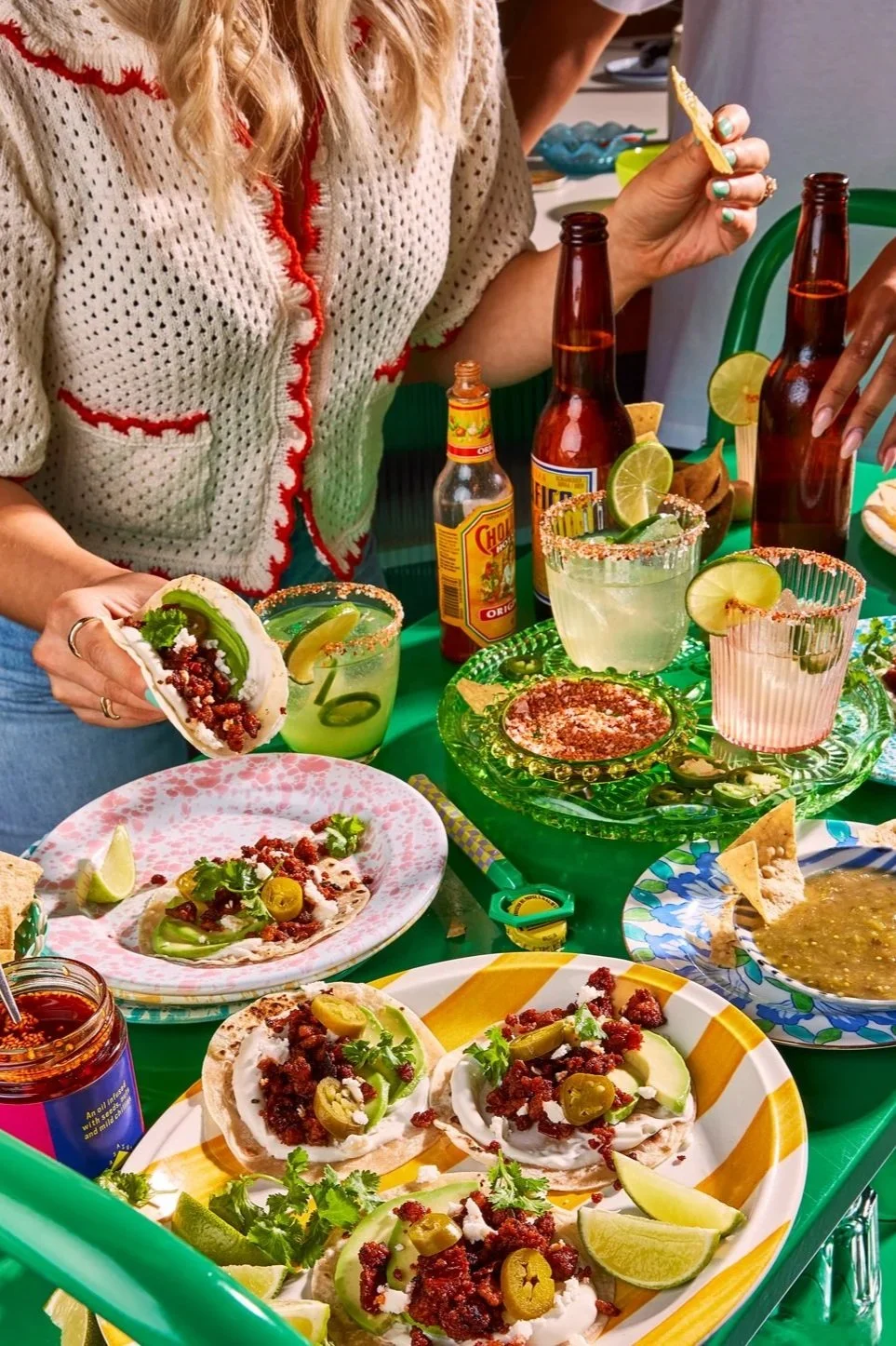 The image shows a table with Mexican food and drinks, including tacos topped with pickled jalapenos, cilantro, and crumbled meat, along with lime wedges. There are several bottles of beer and a jar of hot sauce. A woman is holding a taco and a margar