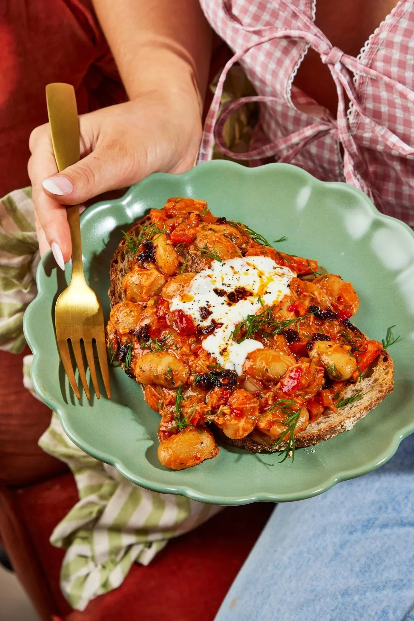 A slice of bread topped with chili, sour cream, and herbs on a green plate, held by a hand with a gold fork in a pink checkered apron.