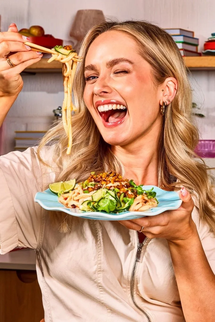 A woman with blonde hair, earrings, and a light-colored jacket is holding a plate of Asian noodle salad garnished with lime and cucumbers. She's smiling and winking while holding chopsticks with noodles above her head.