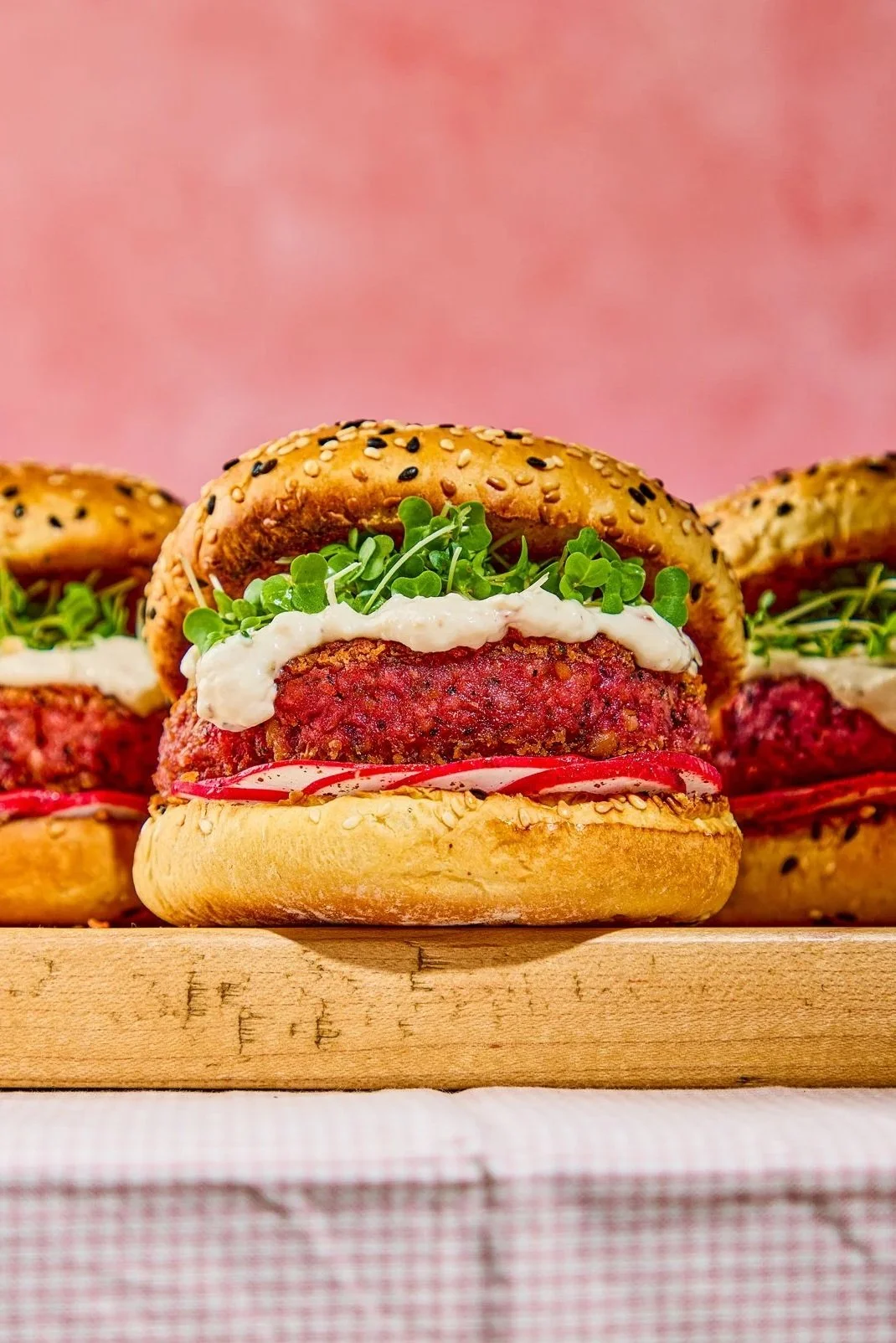 Close-up of a gourmet veggie burger with microgreens, cream sauce, and tomato slices on a bagel bun with sesame seeds, on a wooden surface against a pink background.