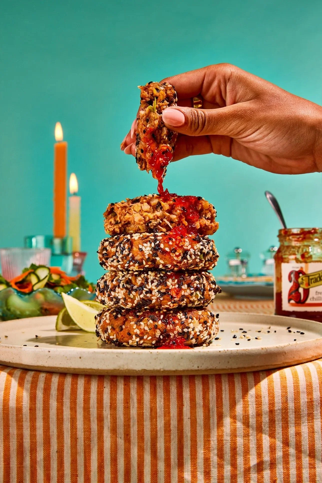 A hand stacking sesame-crusted veggie burger patties with red sauce in between, on a white plate with lemon wedges and a green salad in the background, lit by candles.