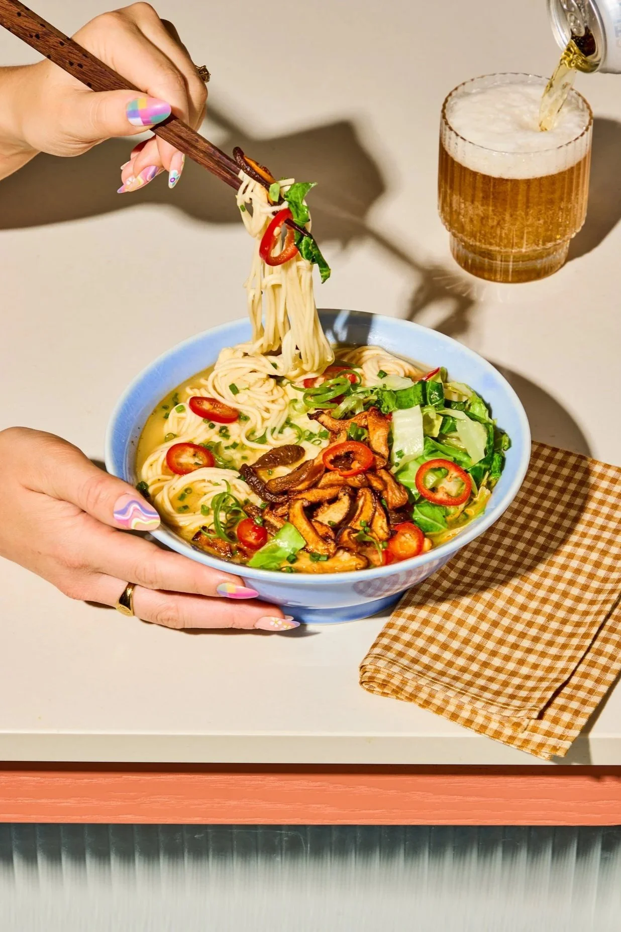 A bowl of ramen with noodles, sliced red chili peppers, chopped green onions, and sautéed mushrooms, with a hand holding chopsticks lifting some noodles, a hand holding the bowl, and beer being poured into a glass in the background.