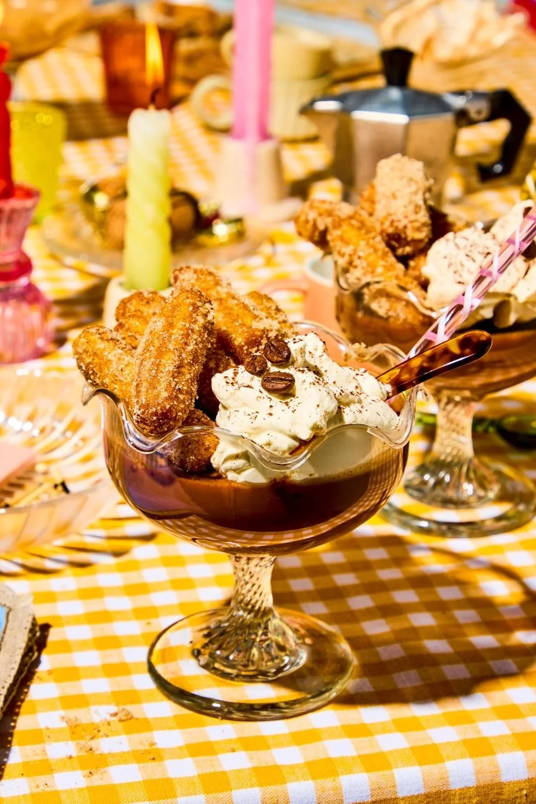 A glass dessert cup filled with whipped cream topped with coffee beans, chocolate syrup, churros, and ice cream, on a yellow and white checkered tablecloth, with a festive background including candles and decorative items.