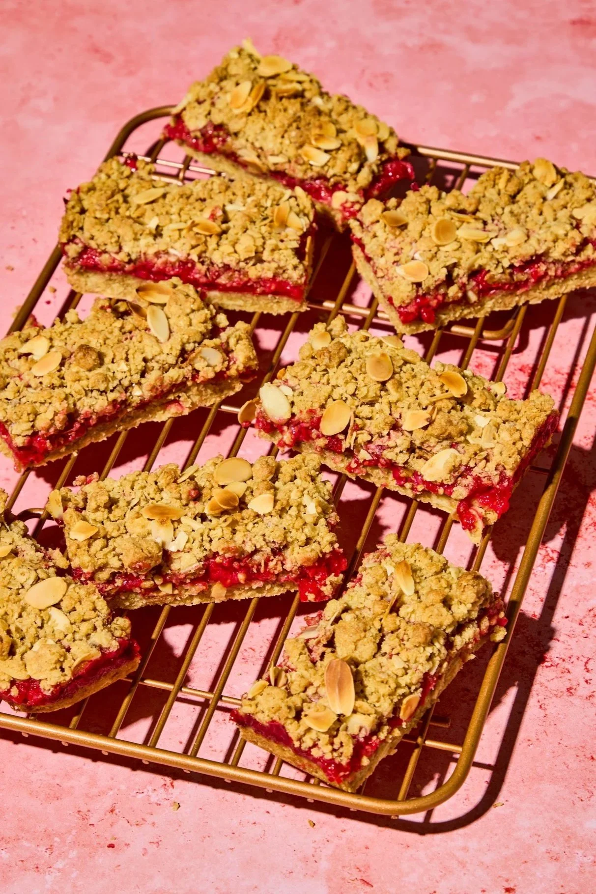 Square berry crumb bars topped with sliced almonds on a metal cooling rack over a pink textured surface.
