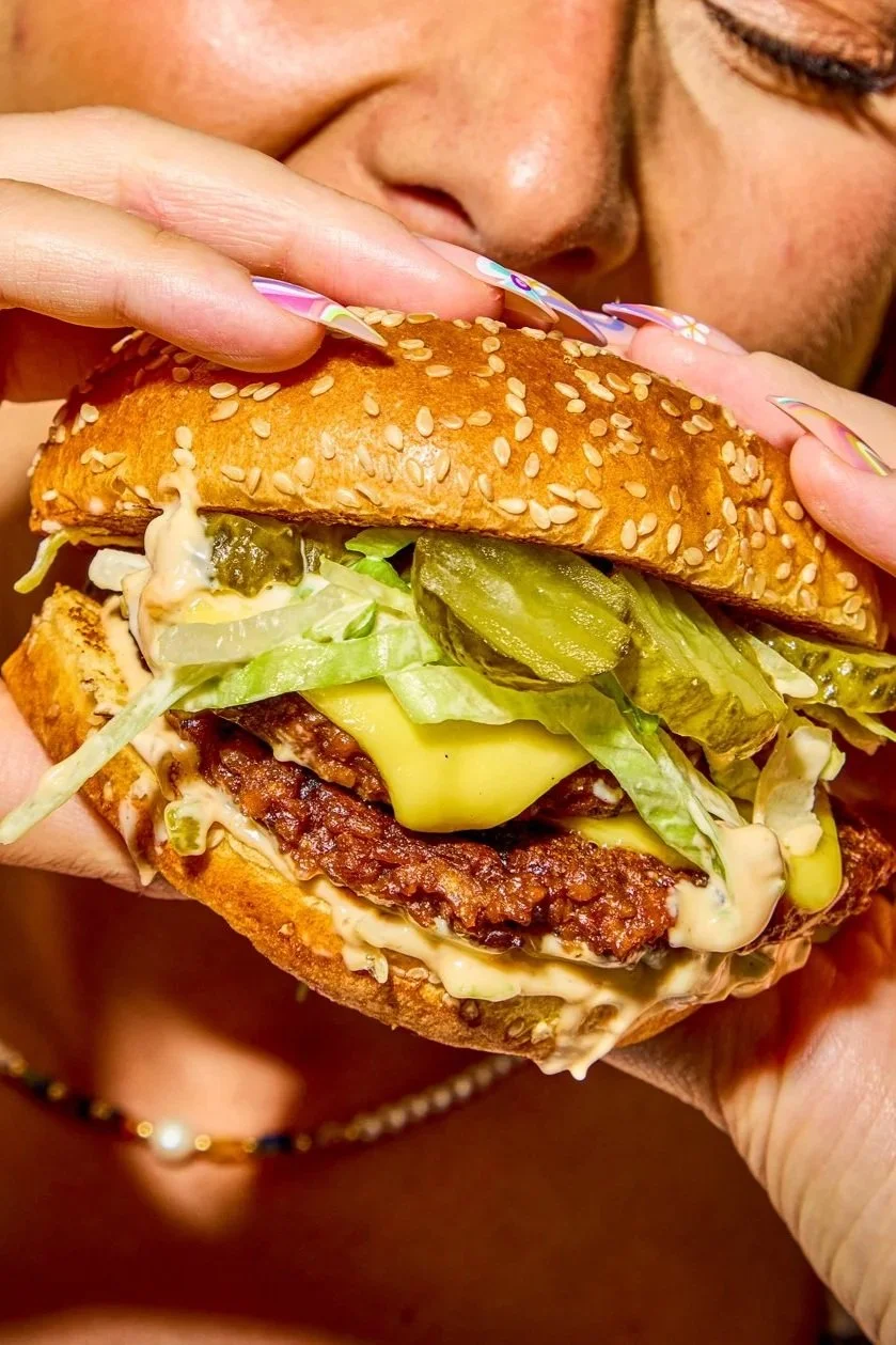 Close-up of a person about to bite into a large burger with sesame seed bun, lettuce, pickles, cheese, and a meat patty.