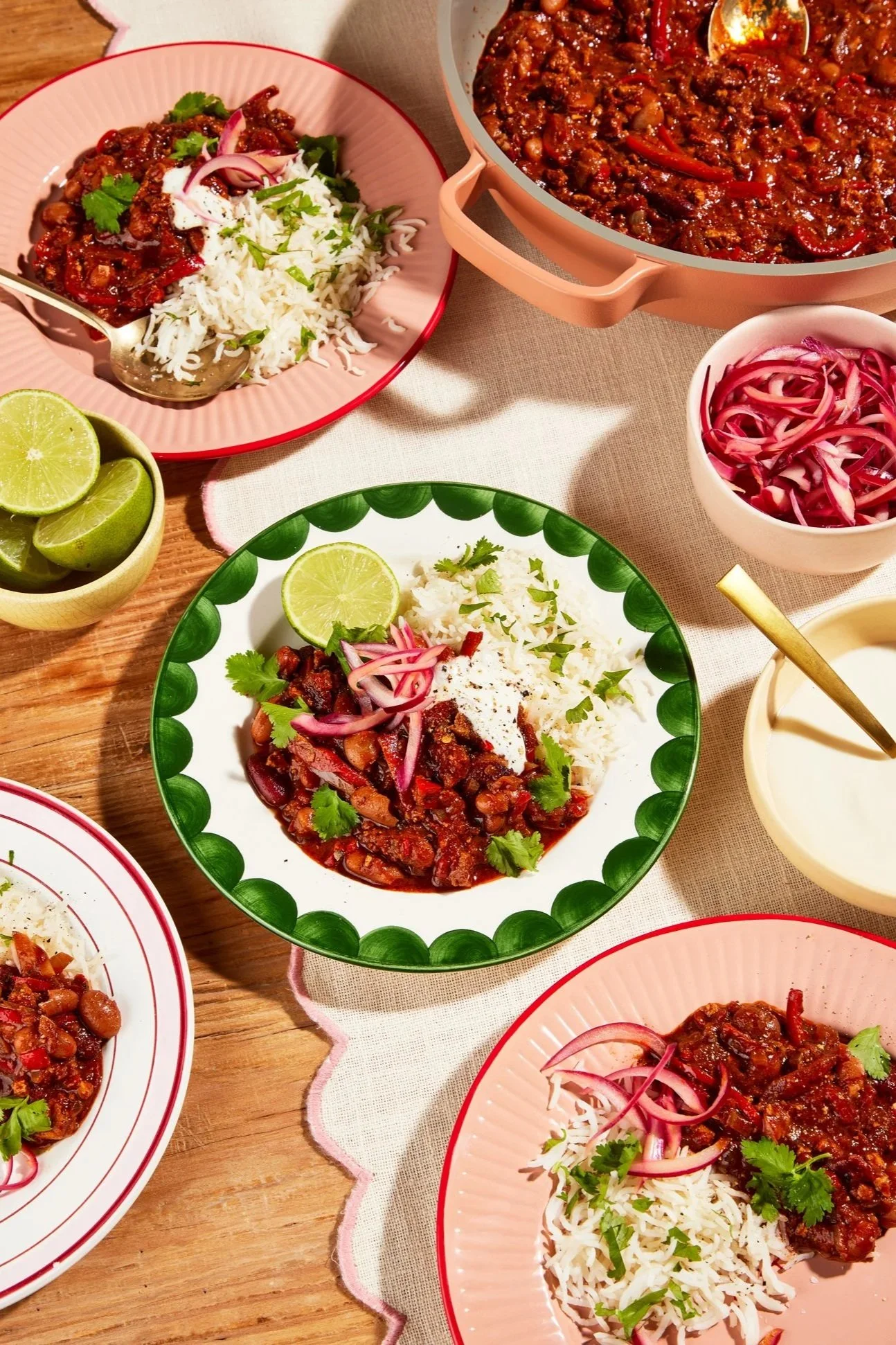 Plate of rice topped with cooked beans, garnished with cilantro and sliced red onions, accompanied by lime wedges, and a bowl of pickled red onions. Several other dishes of similar beans with toppings are visible on a wooden table.