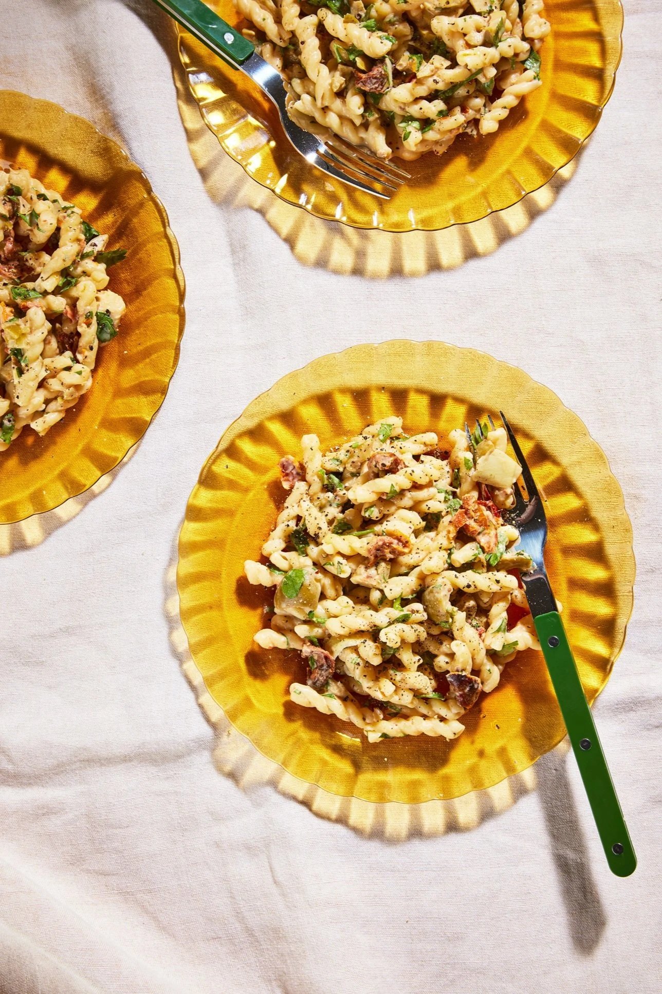 Three yellow glass plates with pasta salad on a white tablecloth, with a green-handled fork on each plate.