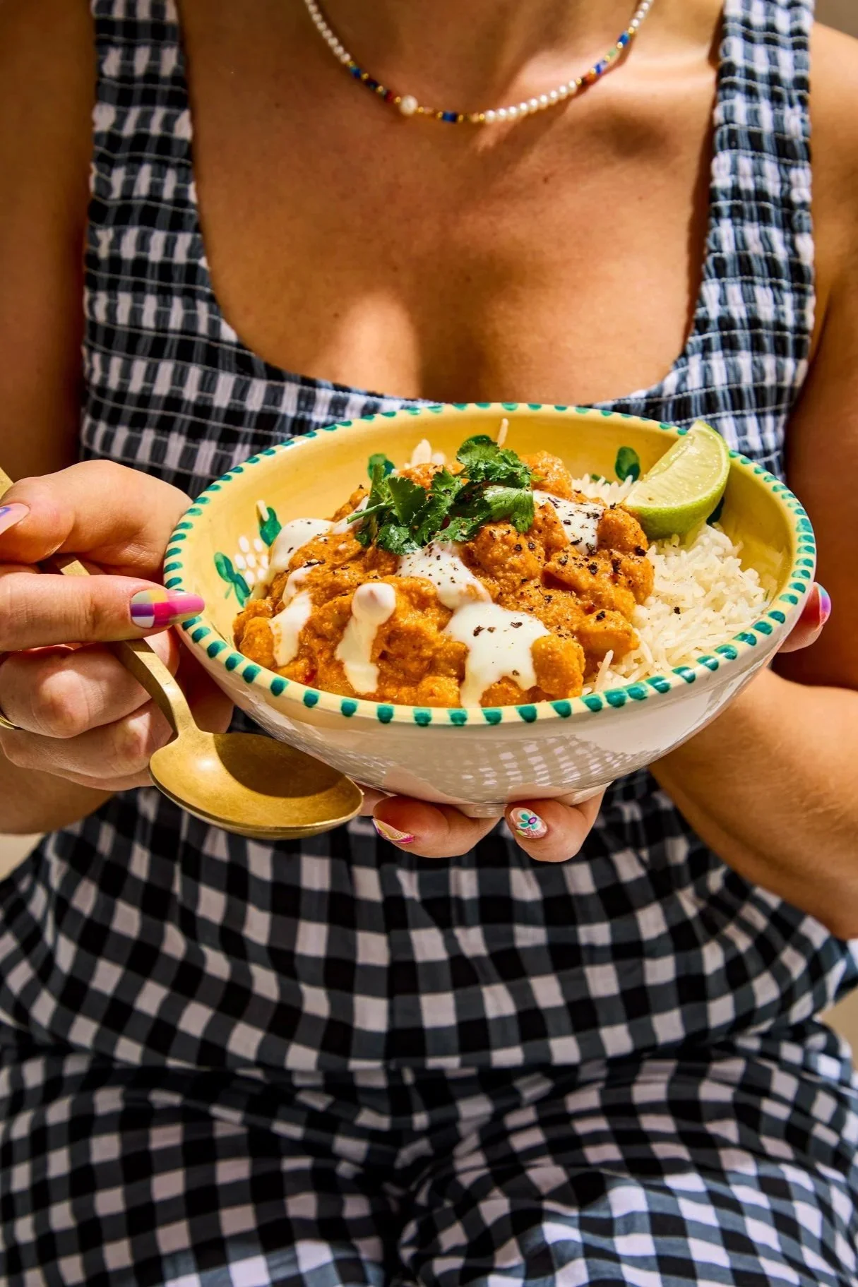 A woman holding a colorful bowl of Indian curry served with rice, garnished with cilantro and a lime wedge, wearing a checkered dress and jewelry.