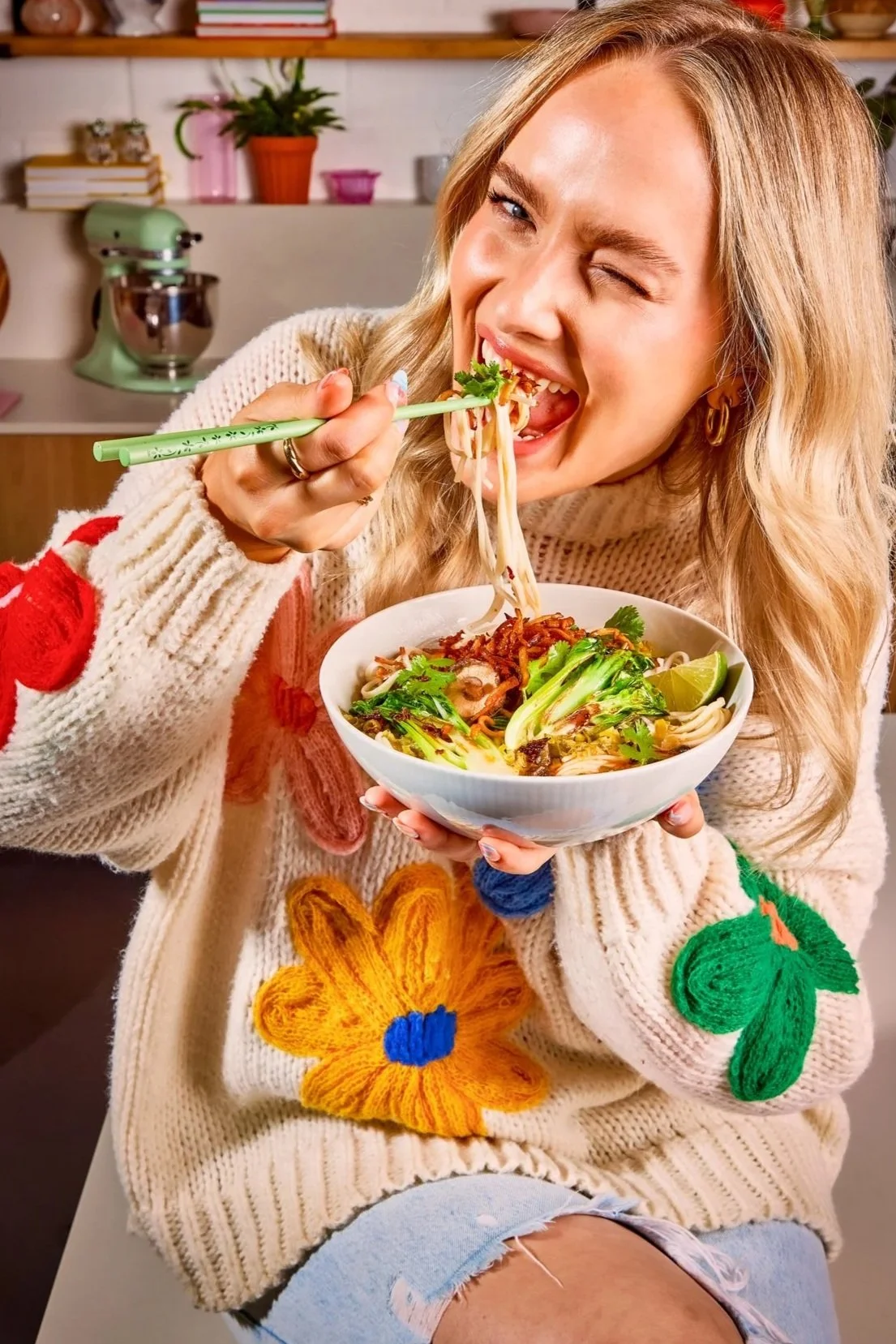 A woman with blonde hair wearing a cream-colored sweater with colorful flower and leaf patches, sitting and eating a bowl of noodle soup with chopsticks in a kitchen.