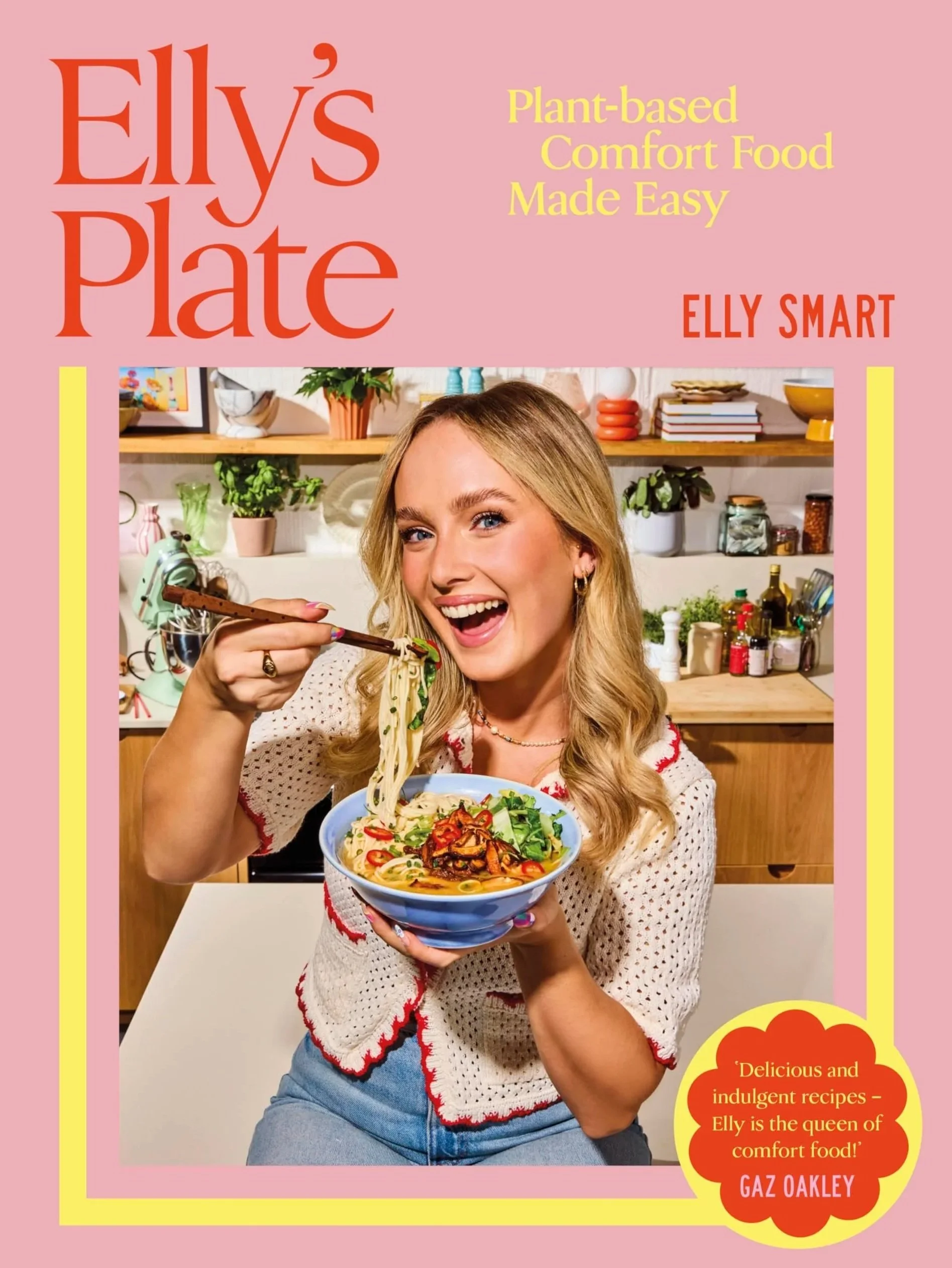 A woman with long blonde hair smiling and holding a bowl of Asian noodle soup garnished with vegetables and chili, standing in a modern kitchen with wooden shelves, plants, and colorful kitchenware.