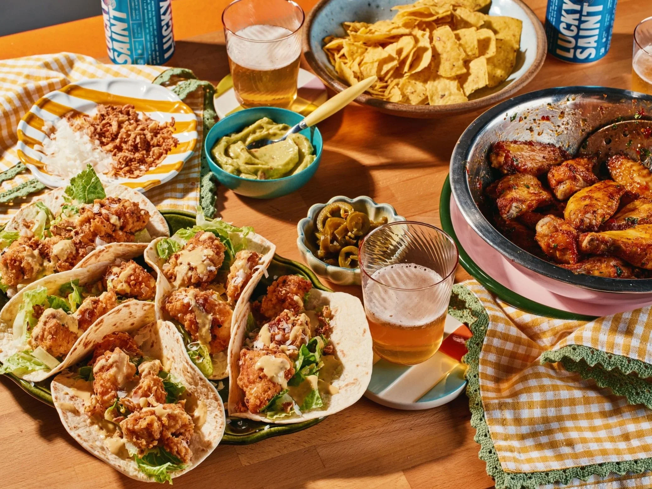 A table filled with tacos, nachos, guacamole, jalapenos, beer, and a variety of Mexican dishes, set on a wooden table with a yellow checkered cloth.