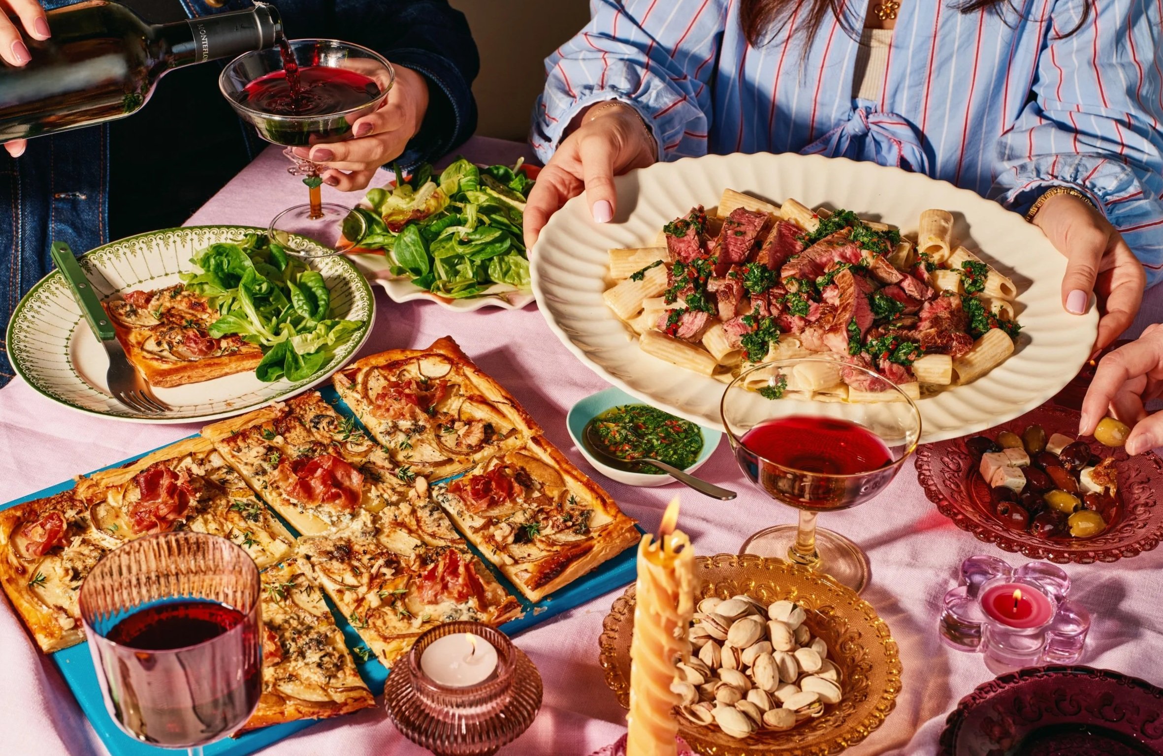 A table filled with various dishes including pizza, pasta, a green salad, a chocolate cake with a candle, and glasses of red wine. Two women are serving food, with one pouring red wine into a glass and the other holding a large plate of pasta topped 