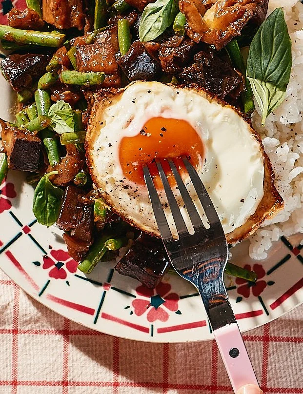 A plate of food with a fried egg on top, surrounded by cooked green beans, diced meat, and rice, on a decorative plate with a fork piercing the egg.