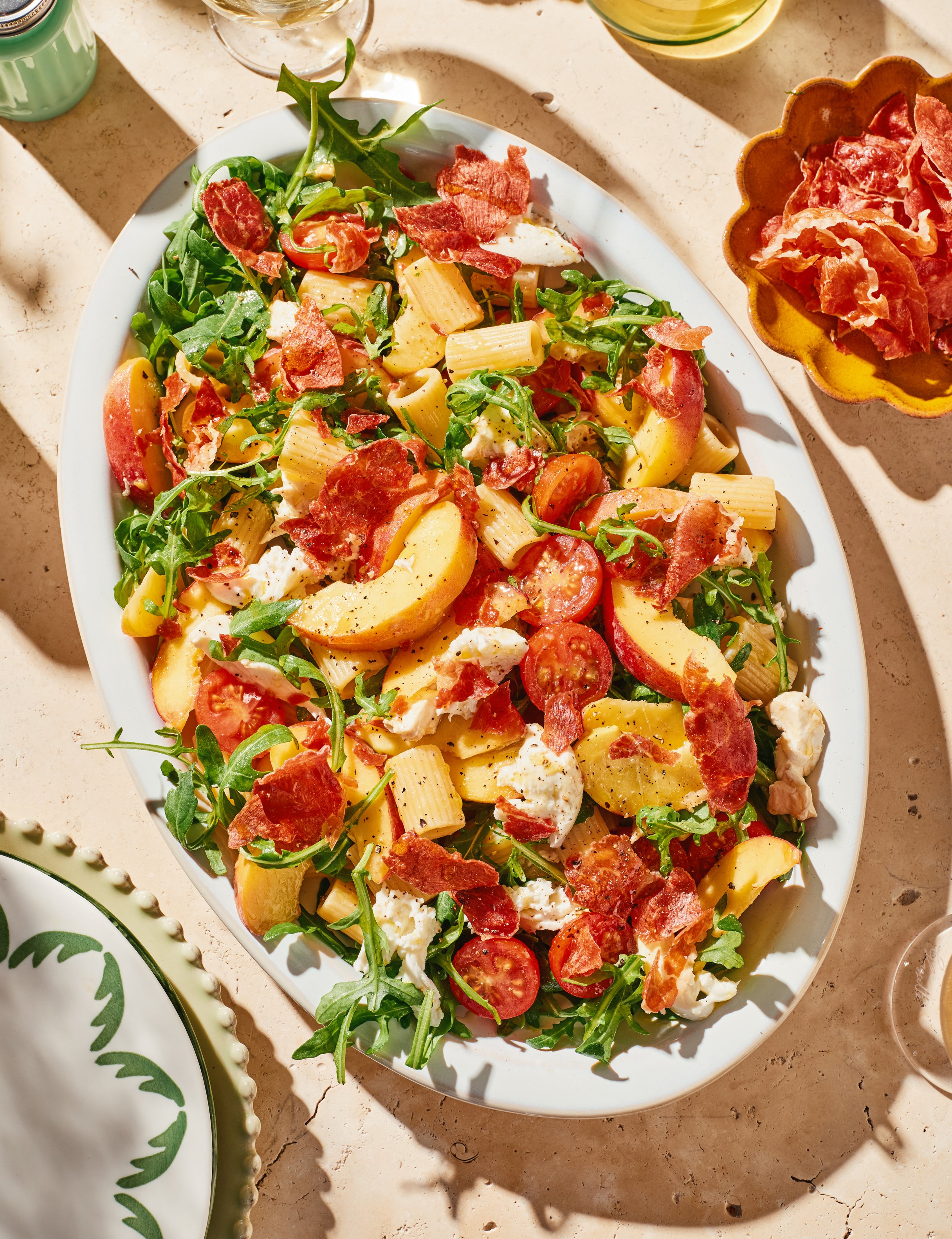 A white oval platter filled with a fresh salad containing arugula, cherry tomatoes, pasta, cheese, and bacon strips, placed on a beige marble surface with shadows.