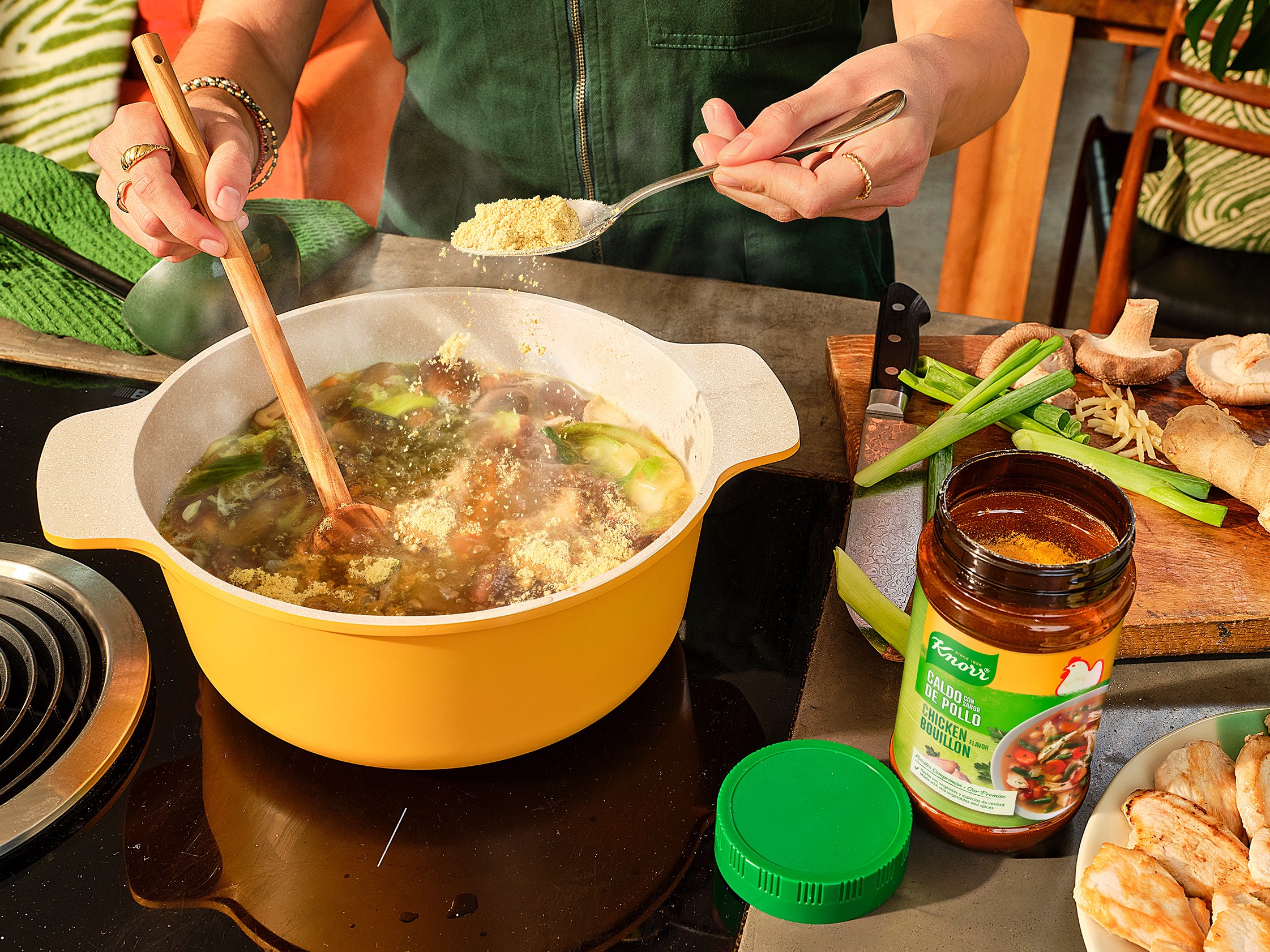 Person scooping chicken bouillon powder over a pot of soup on a kitchen counter, surrounded by various ingredients including mushrooms, green onions, and bread.